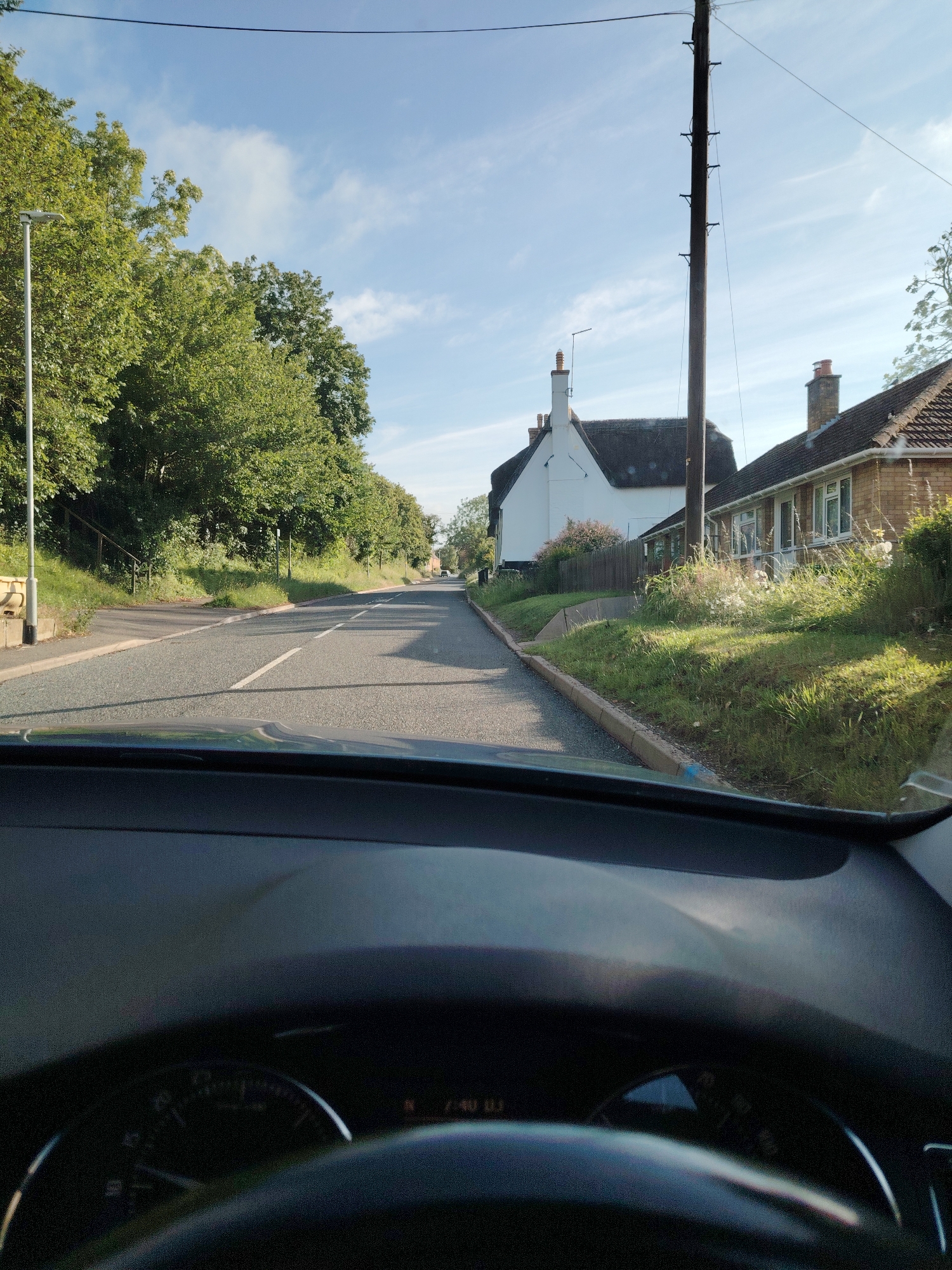 This image, taken from the interior of a vehicle looking forward through the windshield, depicts a rural or village road scene in Spaldwick, Huntingdonshire, United Kingdom. The time displayed on the vehicle's dashboard is approximately "7:40".
The asphalt road, grey in color, extends into the distance with a slight uphill incline and a gentle curve to the right. A single white dashed line marks the center or a lane boundary, while a continuous white line defines the road's right edge.
On the left side of the road, a tall, slender metal streetlamp post stands in the foreground. Beyond it, a steep grassy embankment densely covered with mature green deciduous trees rises alongside the road. A small, lighter-colored utility box or barrier is visible near the road edge at the base of the embankment. Further down, another smaller pole or signpost is present on the bank.
On the right side of the road, a distinct white building with a dark thatched roof is visible, identified as The George Inn. It features a chimney and is partially obscured by foliage at its base. Adjacent to The George Inn, a single-story brick bungalow with a tiled roof is present, featuring multiple windows and a front door. A wooden fence separates these properties from the road. A tall, dark wooden utility pole with several dark crossarms and wires stands prominently between the white thatched building and the brick bungalow. The area between the road and the fence is a grassy verge with scattered wildflowers and taller green vegetation.
The sky above is a clear, light blue with several scattered, thin white clouds. Bright sunlight illuminates the scene, casting shadows, notably from the utility pole towards the left. The bottom portion of the image shows the dark plastic dashboard and a section of the steering wheel of the vehicle, with partial views of the instrument cluster, including a speedometer and the digital time display. No people or animals are visible in the scene.