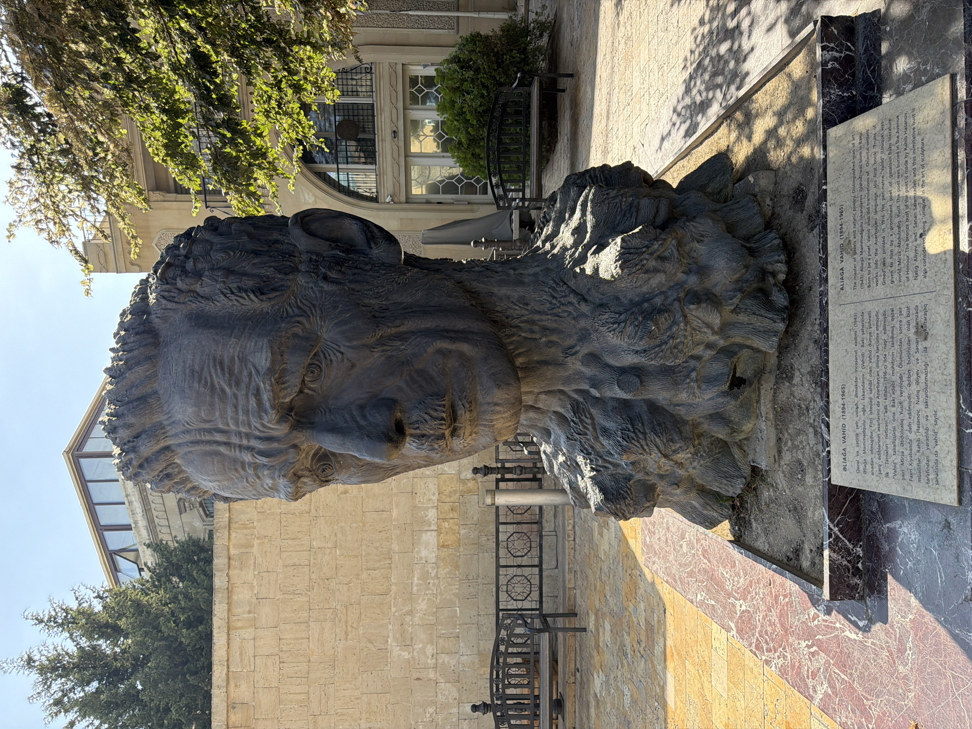 The image shows a large, dark bronze bust of a man in an outdoor setting, likely a public space in Sabail Raion, Azerbaijan. The sculpture depicts the man's head and shoulders, with highly detailed, weathered features. The bust is mounted on a stone pedestal, which is itself set on a marble base. In front of the pedestal is a plaque with text in both Azerbaijani and English, identifying the subject as Aliaga Vahid (1894-1965), a master of satirical poetry and distinguished man of art.
The background features a building with a stone facade and a modern glass-enclosed upper story. Trees with green foliage are visible, casting shadows on the ground, which is paved with stone tiles. Two park benches are positioned in the middle ground, suggesting a park or public square. The sky is clear and blue, indicating a sunny day. The overall scene is well-lit, with strong shadows indicating direct sunlight.