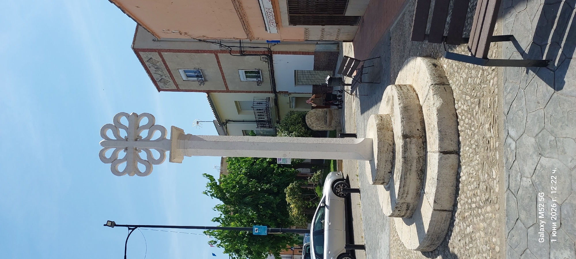The image shows a roundabout in the city of Canals, Spain. A white stone cross is centered in the roundabout with a decorative, clover-like design at the top. The base of the cross has three circular tiers. A silver car is parked nearby. In the background are several buildings, including one with a sign that reads "CARNISSERIA." An elderly person with a walker is seated on a bench in the background. Other benches and a small, stone structure are also present. The sky is clear and blue. A street lamp with wires extends across the top left of the image. A small blue sign with a white figure is visible near the trees. The pavement is textured, with a combination of smooth, geometric patterns and pebbled sections. The photo includes a watermark with "Galaxy M52 5G" and a date of "1 June 2026."