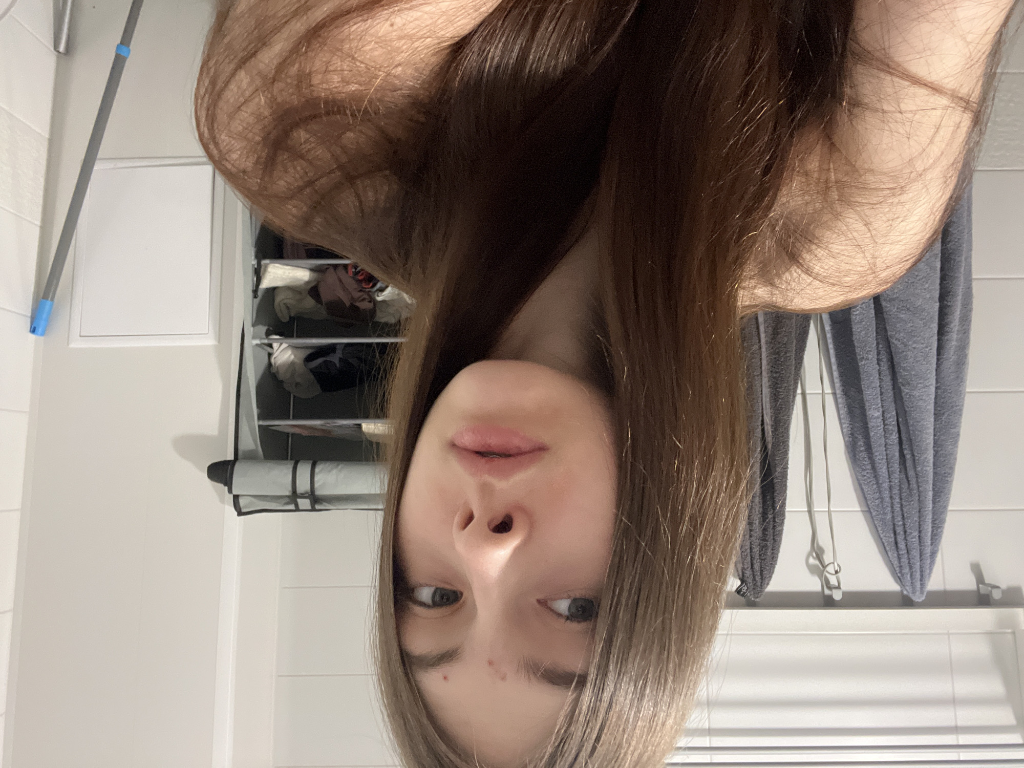 An inverted portrait features a young individual, likely female, with long, straight brown hair cascading upwards from the bottom of the frame. Her face is visible in the lower central part of the image, with light-colored eyes looking directly towards the viewer. Her lips are slightly parted, and her expression is neutral. A couple of small blemishes are noticeable on her chin/forehead area in the inverted view.
The background, also inverted, reveals an indoor setting, strongly suggesting a bathroom or utility room in Saint Petersburg, Russia. White tiled walls are prominent throughout. To the left, a gray mop or cleaning tool with a blue handle cap leans against the wall. A rectangular white panel or door is also visible on the wall. To the right, a multi-tiered storage unit or shelving system contains various folded items, possibly clothes or towels. Further to the right, a gray, fluffy towel or piece of fabric hangs from a white railing or towel rack. The lighting is even, suggesting an indoor light source. No discernible text is visible in the image.
