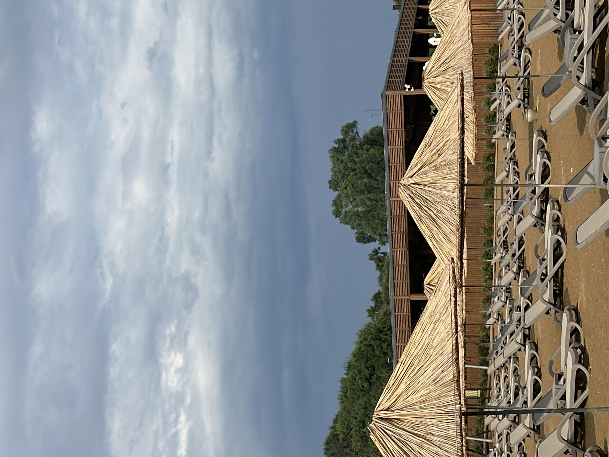 This image depicts a beach resort area in Manavgat, Turkey, likely on a cloudy day. In the foreground, rows of empty lounge chairs are arranged on a sandy surface, covered by large, thatched umbrellas. Behind the chairs and umbrellas, a wooden structure resembling a building or pavilion with horizontal slats runs across the scene. Further in the background, dense green trees and foliage are visible. The sky above is overcast with a mix of light and dark gray clouds. The scene appears to be devoid of people, suggesting it might be early morning, late evening, or off-season.