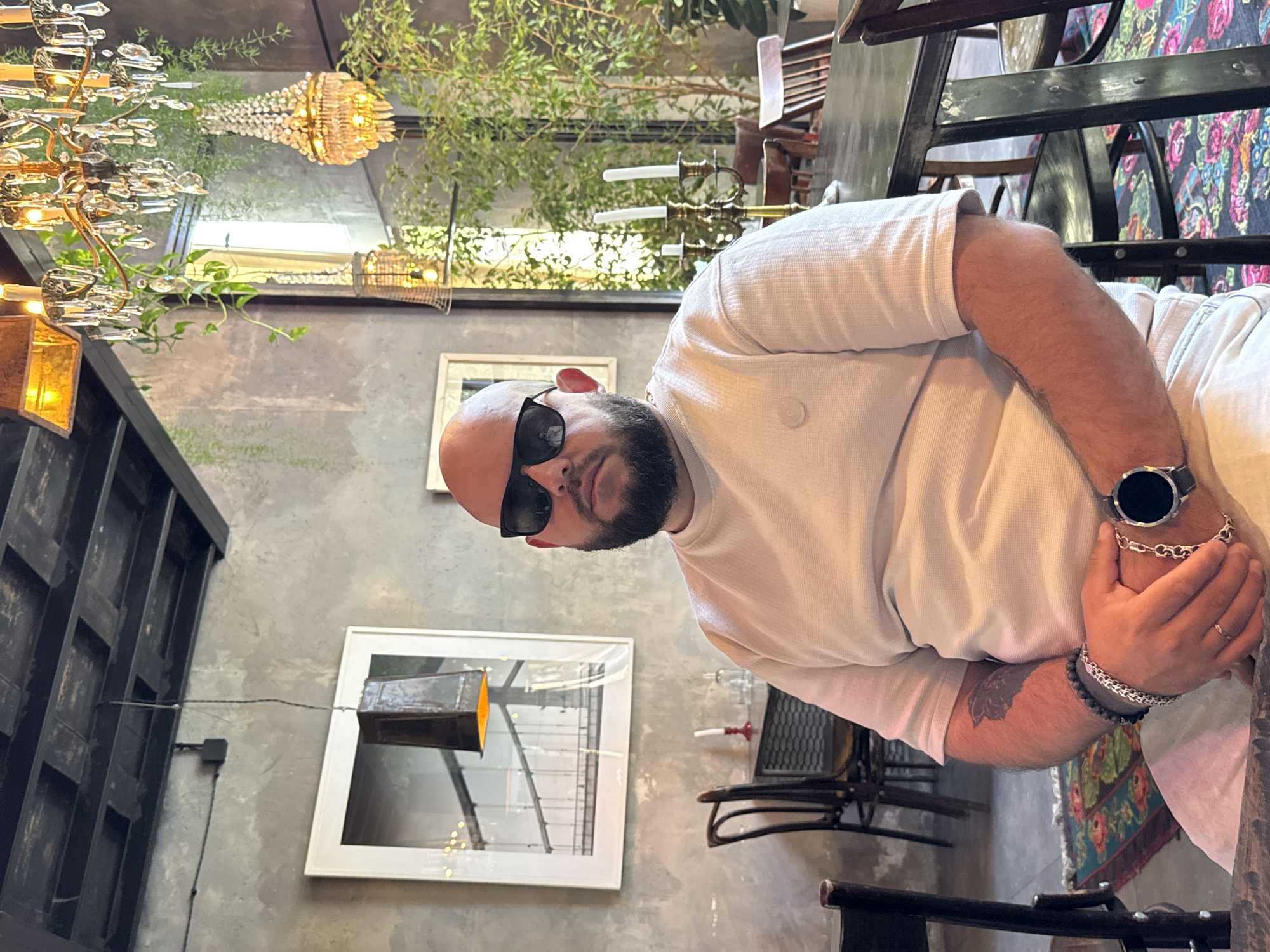 A man with a shaved head and sunglasses, wearing a white waffle knit shirt, sits at a table in what appears to be a restaurant. He is wearing a watch and bracelets on his left wrist. The background includes gray walls adorned with framed pictures and decorative lighting fixtures, including chandeliers. Green plants are also visible, adding to the ambiance of the establishment. The location appears to be a restaurant with an interior design reflecting a specific style or aesthetic. The location is not explicitly identified as Krasnaya Polyana, Russia, but that information was included in the request.
