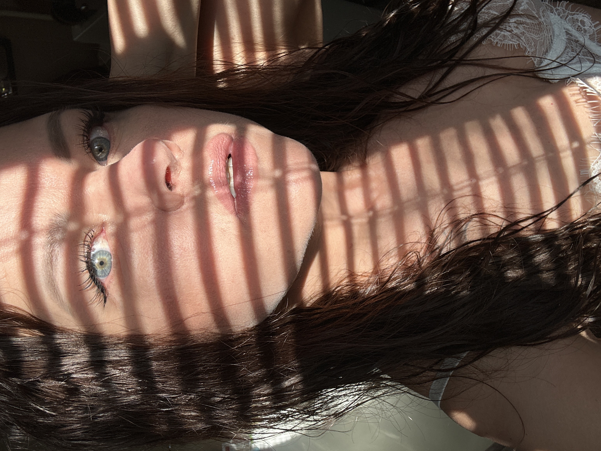 A woman with long, dark hair lies on her back, her face illuminated by sunlight streaming through blinds. The light creates strong shadows across her face, revealing clear blue eyes, full lips, and fair skin. She is wearing a white lace garment. The location appears to be indoors, possibly a bedroom. The image does not provide enough information to determine if the location is Chaikovsky, Russia.