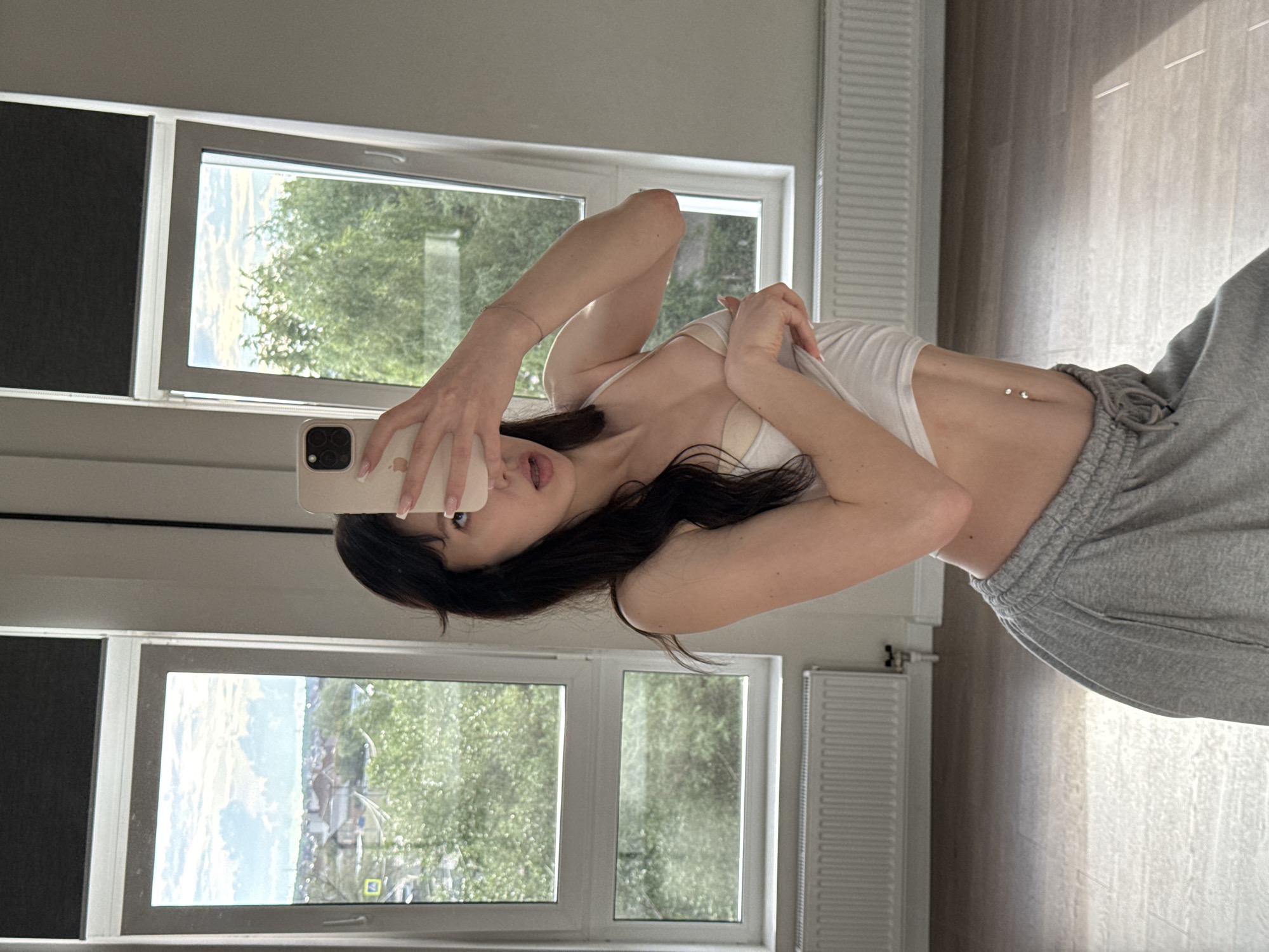The image shows a young woman with long, dark hair, taking a selfie in a room with large windows. She is wearing a white crop top and grey sweatpants. Her left arm is raised, holding a phone, and her right arm is bent, with her hand near her face. She has a belly button piercing with two small jewels.
Through the windows, a green, leafy landscape is visible, suggesting it is daytime and likely a pleasant season. The room appears to be a modern living space, with radiators visible along the wall. The lighting suggests it is daytime, possibly late morning or afternoon, with sunlight filtering in, casting shadows on the wall.
The woman's expression is playful, with her mouth slightly open and her tongue peeking out. The overall mood is casual and relaxed. The image appears to have been taken indoors, possibly in a home in Zakharovo, Russia, given the location context provided. No visible text is present in the image.
