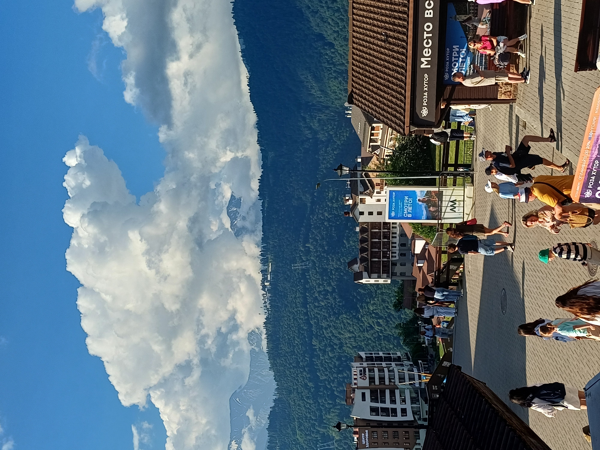 This image depicts a sunny day in Estosadok, Russia, a resort town in the Krasnodar Krai region. The scene is set in a pedestrian area with paved ground and buildings in the background. In the foreground, several people are walking and standing. To the left, a woman with a backpack walks away from the camera. To her right, a group of people, including children, are walking towards the camera. Further into the scene, more individuals are seen milling about.
The background is dominated by the lush green slopes of the mountains, characteristic of the Caucasus region. The mountains are partially shrouded in dramatic clouds, with snow-capped peaks visible in the distance. Several multi-story buildings, appearing to be hotels or apartments, are nestled into the lower slopes. One building on the left has the text "ЦЕНТР ЗДОРОВЬЯ" visible. A tall blue billboard with Russian text, likely an advertisement, stands near the center-left.
A prominent building with a dark brown tiled roof is situated in the right mid-ground, with a sign above its entrance reading "РОЗА ХУТОР Место вс". This indicates the location is part of the Rosa Khutor resort. People are visible sitting on a bench in front of this building. The overall atmosphere suggests a lively tourist destination, likely during the summer season, with visitors enjoying the scenic mountain landscape. The lighting suggests it is late afternoon due to the long shadows cast on the ground.