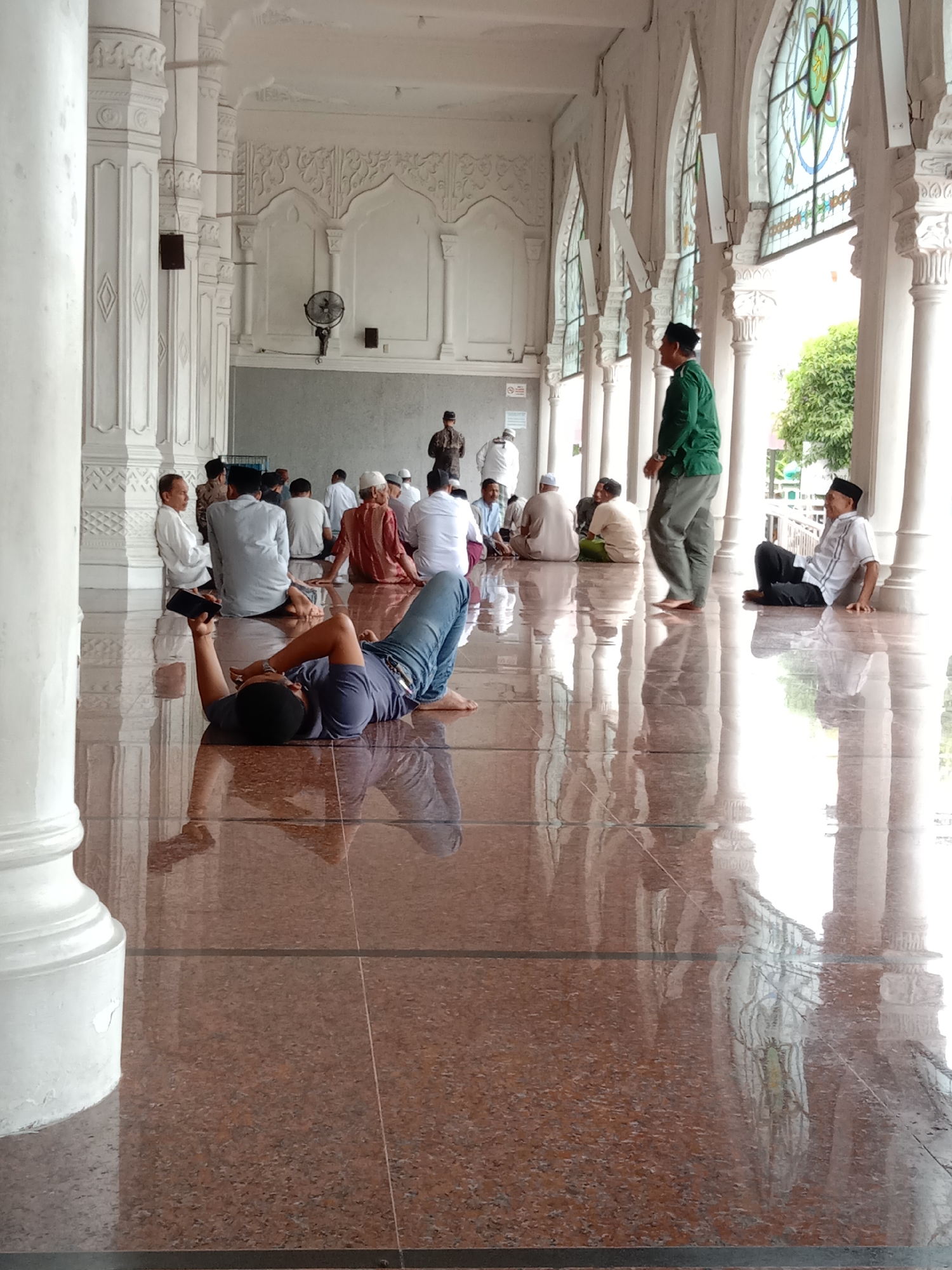 The image captures a scene within the Islamic Center Lhokseumawe mosque, located in Lhokseumawe, Indonesia. The interior space is characterized by a polished, reflective brown tiled floor and white ornate pillars that frame arched windows with colorful glasswork.
A group of men are seated on the floor in the background, some wearing traditional Muslim attire, including white caps. A man in a green shirt and grey pants walks among the seated men. In the foreground, a man lies on his back, holding a black object, possibly a phone, above his head. He is wearing a blue t-shirt and jeans. His body is angled diagonally across the foreground of the image. The scene suggests a moment of rest or leisure within the mosque.