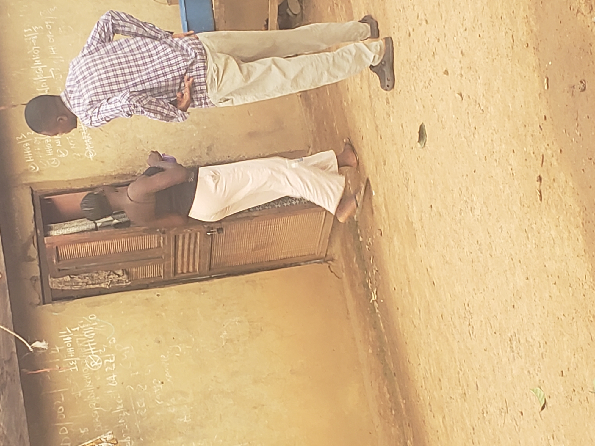 The image depicts two young Black boys in what appears to be an outdoor or semi-outdoor classroom setting. The ground is made of dirt, and there is a rough-hewn wooden structure in the background, possibly a cabinet or doorway with slatted doors. One boy, wearing a checkered shirt and light-colored trousers, is standing and facing away from the camera, with his hands behind his back. The other boy, wearing a dark top and white trousers, is kneeling or crouching in front of a dirt wall, facing towards the same wall where writing is visible.
The wall has chalk writing on it, including numbers, tally marks, and some characters that are difficult to decipher clearly. Some of the visible text appears to be mathematical or educational in nature, with phrases like "16/10/2021", "3/16", and numbers such as "22" and "4". There are also tally marks indicating counts.
The overall scene suggests a casual educational environment, possibly in a rural or less developed area. The lighting suggests it is daytime, and the setting appears to be relatively dry, given the dirt ground. The expressions or emotions of the subjects are not clearly discernible due to their poses and the image quality. The presence of educational writing on the wall strongly indicates a learning activity.