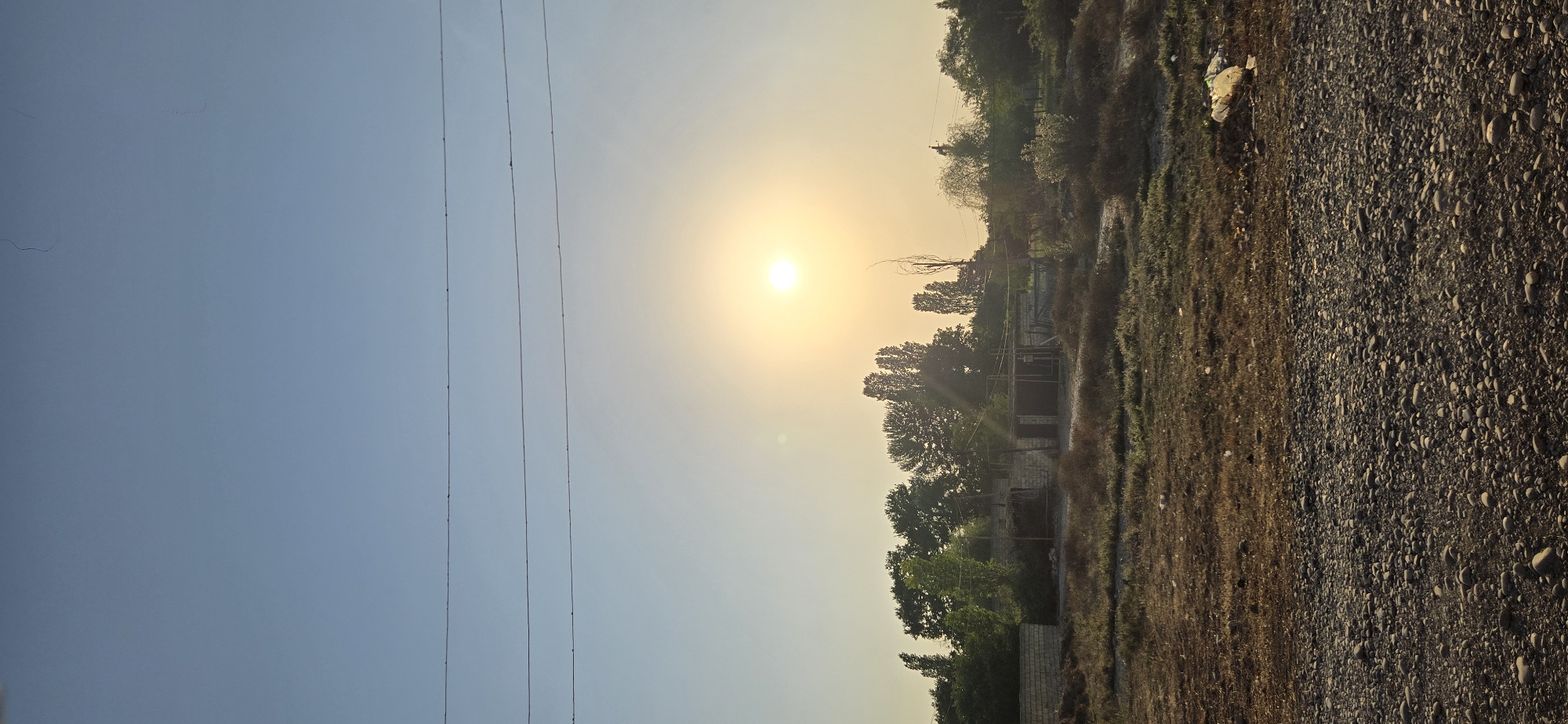 The image captures a rural landscape in the city of Tel, Azerbaijan, during what appears to be early morning or late afternoon. The sky is a clear, pale blue, transitioning to a hazy yellow-orange near the horizon where the sun is visible as a bright, diffused orb. Three parallel power lines stretch horizontally across the upper portion of the frame, with small insulators dotting their lengths.
The middle ground features a collection of low-lying structures, possibly sheds or unfinished buildings, constructed with light-colored brick and topped with wooden framing and corrugated metal roofs. These structures are nestled amongst a dense growth of trees with varying shades of green foliage. Some of the trees are tall and slender, while others are broader and more rounded. The ground in front of these structures is covered with dry, patchy vegetation, interspersed with soil and small stones.
The foreground is dominated by a rough, gravelly surface, densely packed with small to medium-sized pebbles and stones of various grey and brown hues. To the right of the foreground, on the edge of the grassy area, a white plastic bag is visible, suggesting the presence of litter. The overall impression is one of a somewhat undeveloped or rural area, possibly on the outskirts of a settlement, with signs of both natural elements and human construction. There are no people or animals visible in the scene.