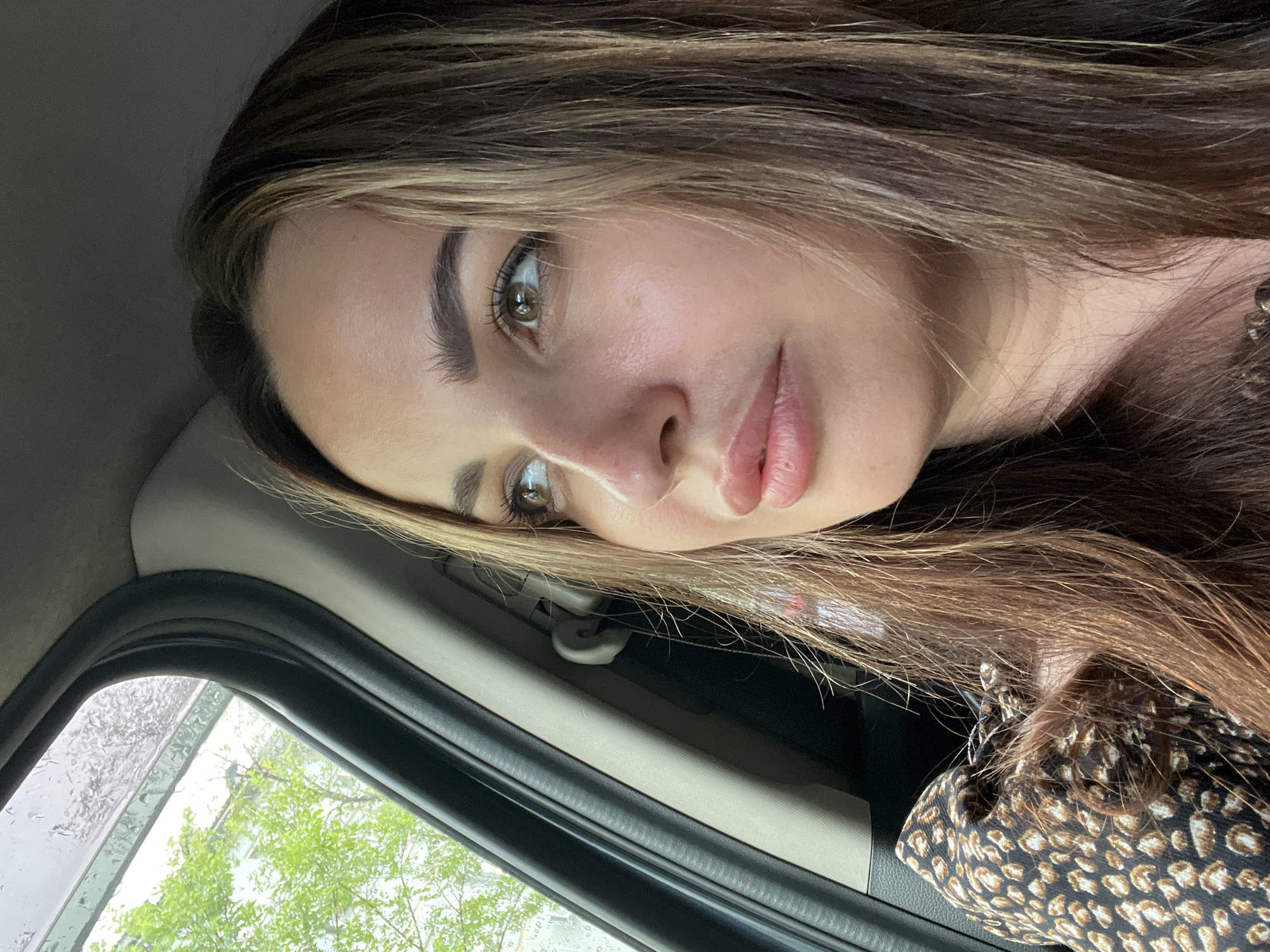 A young woman with long, wavy brown hair is looking out the window of a car. She is wearing a leopard print top. The window is partially open, and the glass has raindrops on it. Outside the window, green trees and a building are visible. The interior of the car, including the door frame and a seatbelt, is also visible.