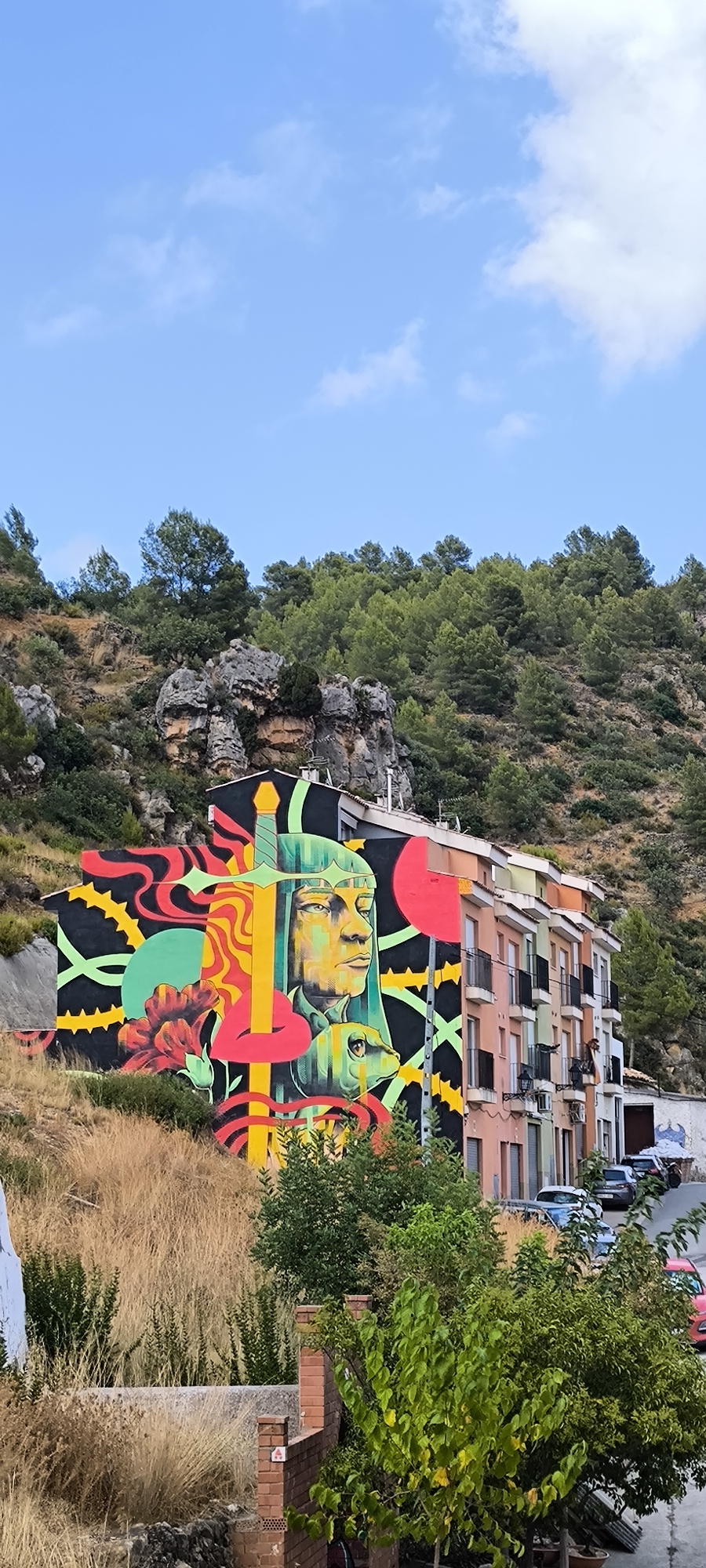 The image shows a building with a large mural on its side. The mural depicts a stylized human face with a headdress, a sword, a flower, and abstract shapes in vibrant colors like yellow, red, green, and black. The building itself is multi-story with a row of balconies, and it is painted in various pastel colors. It is located in a hillside environment with trees and rocks visible in the background. In front of the building, there is a street with several parked cars. The sky is blue with some scattered clouds. This mural is located in Fanzara, Spain, which is known for its street art.