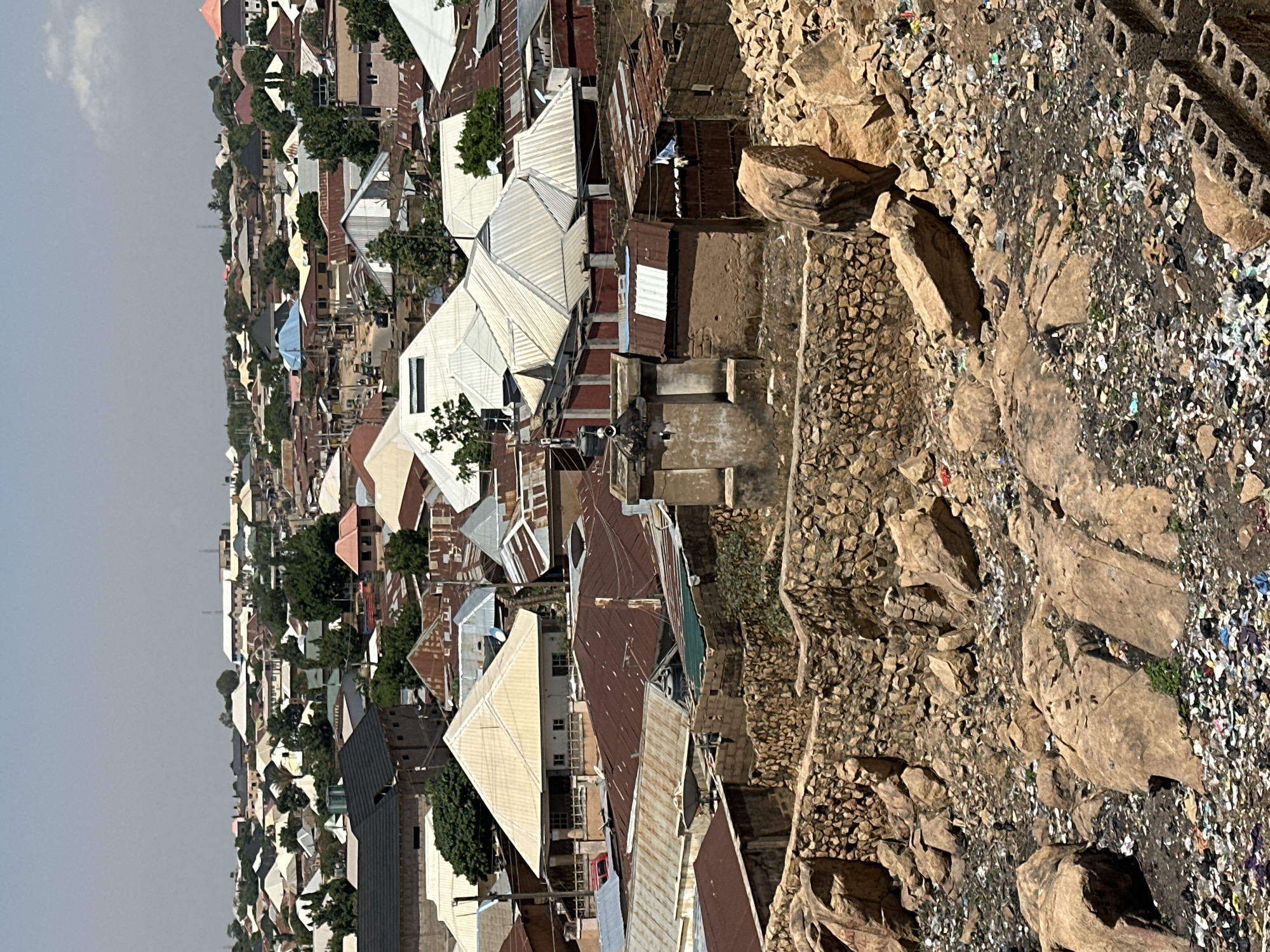 The image presents an elevated view of a dense residential area in Jos, Nigeria, adjacent to a rocky hillside. The built environment is characterized by closely packed houses with a variety of roof types, including corrugated metal and painted surfaces. Many roofs appear aged and weathered. Vegetation, consisting of trees and shrubs, is interspersed among the houses.
The foreground features a steep, rocky slope with loose debris, including what appears to be trash. Stone retaining walls are constructed along the hillside to manage the terrain. The background shows the continuation of the residential area receding into the distance under a somewhat hazy sky. There are no people visible in the frame. The overall impression is that of a densely populated urban area bordering a natural, less managed environment.