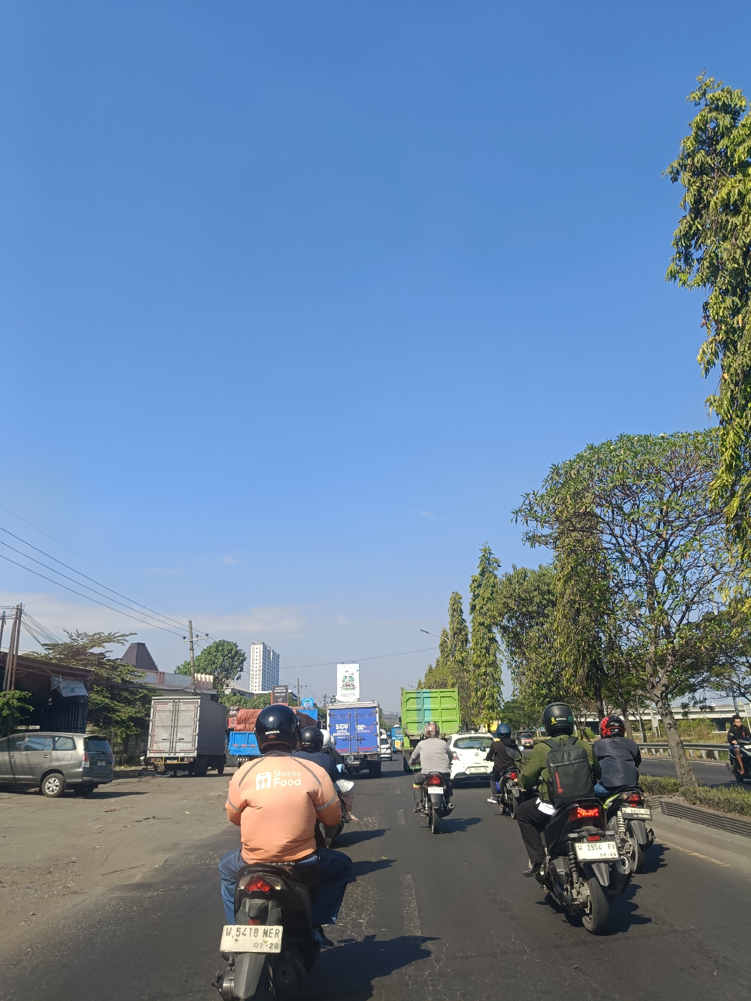 This is a photograph of a street scene in Dahanrejo, Indonesia, on a clear, sunny day. The image is taken from the perspective of someone riding on the back of a motorcycle, looking forward.
In the foreground, a person wearing an orange shirt with "Shopee Food" branding is riding a motorcycle, facing away from the camera. They are wearing blue jeans and a black helmet. The motorcycle has a license plate that reads "W 5418 NER 09-20". Another motorcycle is partially visible in front of them.
The street appears to be a multi-lane road with traffic. Several other motorcycles are visible in front, including one with a rider wearing a green jacket and a backpack, and another with a rider wearing a dark blue jacket and a red helmet. Further ahead, there are cars and large trucks, including a grey truck with an open trailer and a green truck. A silver SUV is parked on the left side of the road.
The environment includes buildings and vegetation. On the left, there are some structures that appear to be shops or warehouses, along with trees and utility poles with power lines. In the background, a tall, modern building can be seen. On the right side of the road, there are trees lining the street and a bridge or overpass. The sky is clear blue.