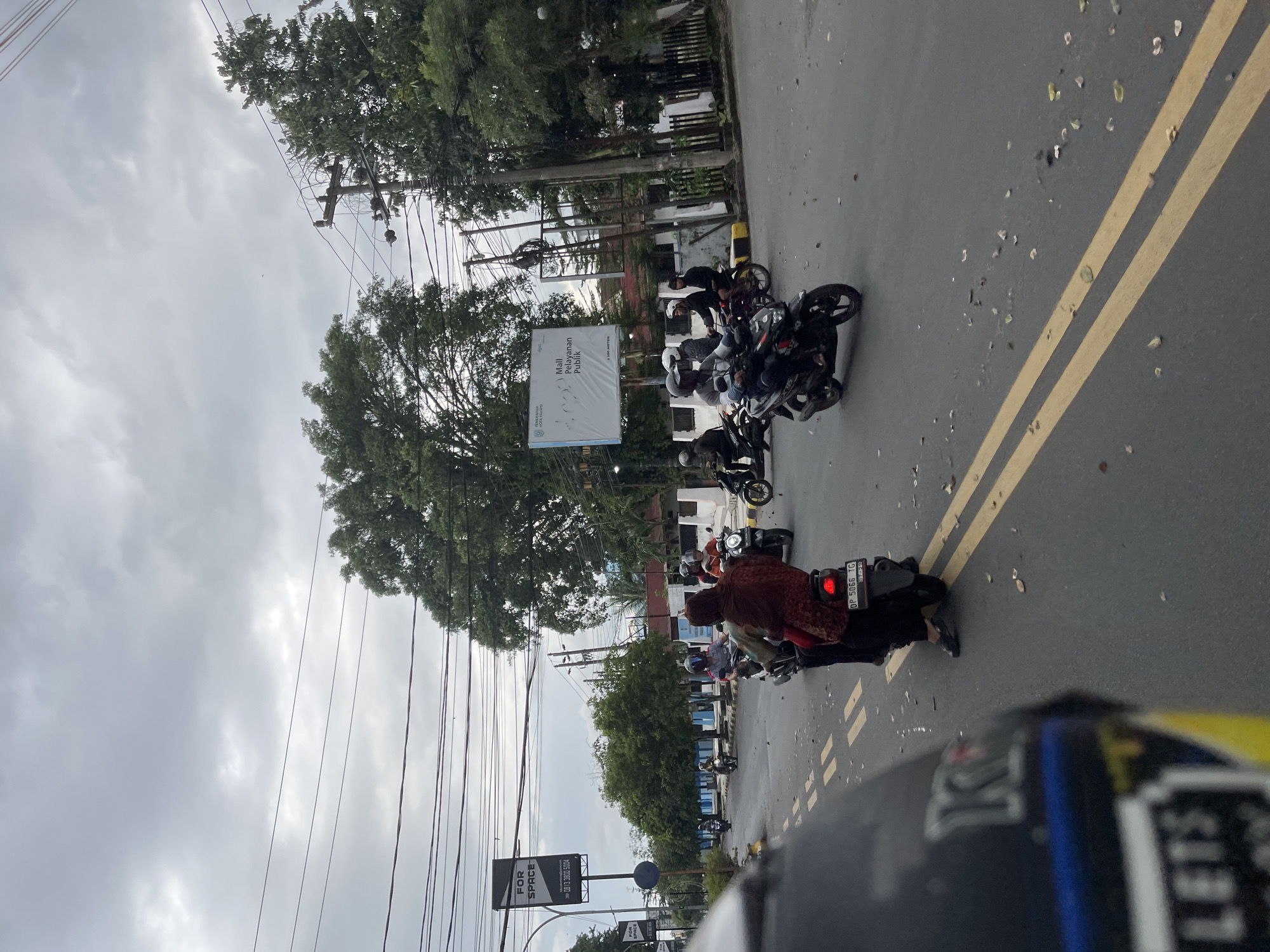 A group of people on motorcycles are gathered on the side of a wet asphalt road. The sky is overcast, suggesting it is a cloudy day, possibly after rain. The road has a double yellow line marking. Trees line the side of the road, and power lines are visible overhead. There are several motorcycles parked and some people are seated on them, wearing helmets and casual clothing. One person in the foreground is wearing a patterned maroon outer garment. A sign with text is visible on a pole behind some of the motorcycles. Another sign further down the road has text that reads "FOR SPACE". The license plate on one of the motorcycles is visible and reads "DP 5066 TC". The overall scene appears to be a casual gathering or pause in travel. The presence of multiple motorcycles and people suggests a social or practical reason for their assembly. The wet road and cloudy sky indicate recent or ongoing precipitation.