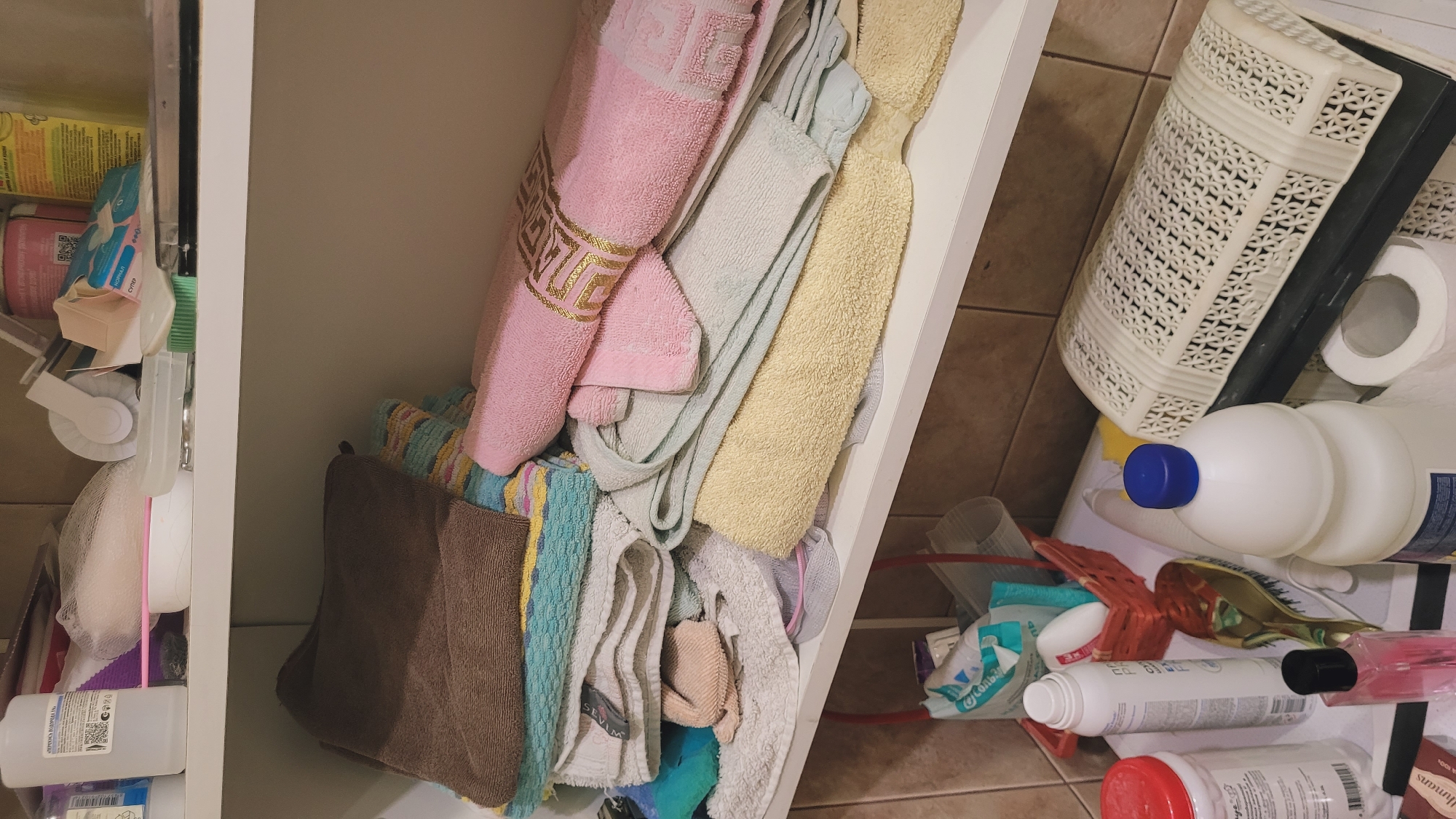 The image shows a close-up view of a shelf in a bathroom or storage area. The shelf is filled with folded towels of various colors, including brown, pink, yellow, blue, and white. Some towels are neatly stacked, while others are piled more haphazardly. On the shelf below, there are several bottles and containers, including a large white bottle with a blue cap, a white spray bottle, and a small pink bottle. A red basket containing some items is also visible on the lower shelf. The background consists of tiled walls. The lighting appears to be artificial, casting shadows on the objects. The scene depicts an organized, albeit somewhat cluttered, storage space.
