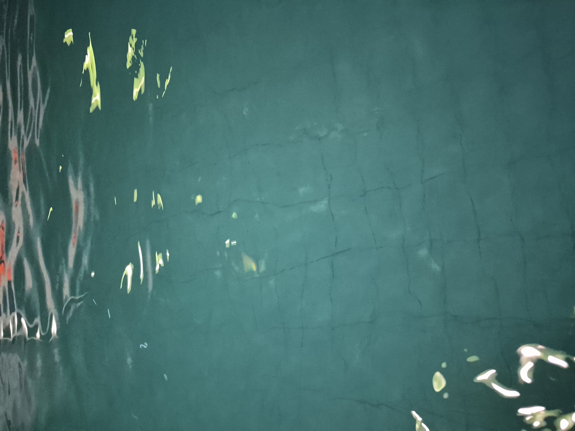 The image depicts the surface of a body of water, likely a river or canal, in Tobolsk, Russia. Reflections of objects (possibly buildings and foliage) are visible in the water, causing distorted shapes and light patterns. The water's color is a dark turquoise-green. A grid-like pattern is faintly visible under the surface, possibly indicating the presence of paving stones or another structured surface below the water.