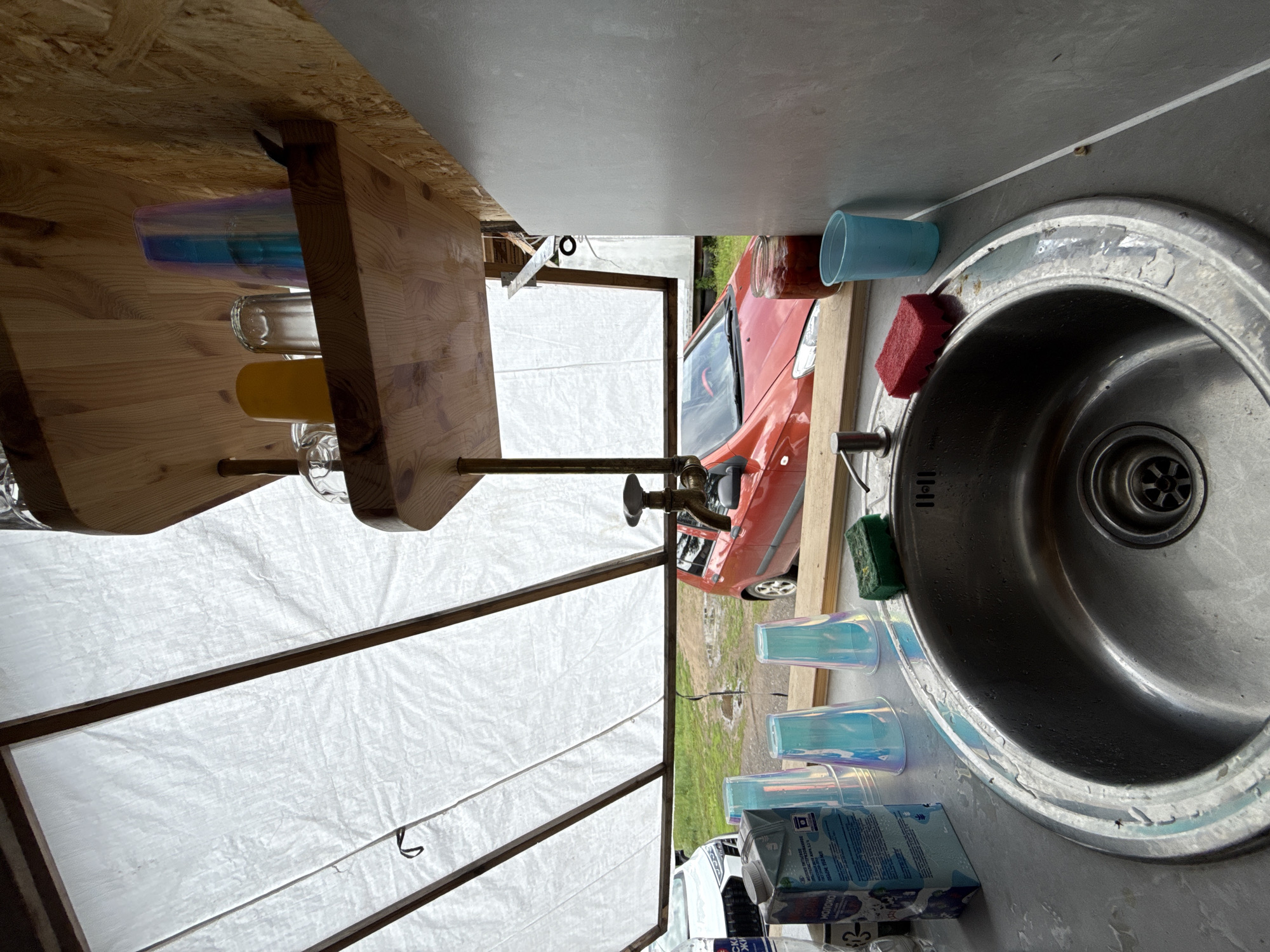 The image depicts an outdoor scene with a sink area in the foreground, set against a backdrop of cars and a large white tarp. A stainless steel sink is prominent, with a faucet, a green sponge on its left, and a red sponge on its right. To the left of the sink, a carton of milk with Russian text is visible. Behind the sink, iridescent plastic cups are arranged, along with a jar containing red contents. A red car is parked in the middle ground, with a white car partially visible on the left. The scene appears to be in an open area, possibly a campsite or a temporary setup, with a wooden shelf holding glassware and other items above the sink area. The presence of the cars suggests an urban or semi-urban environment. The overall impression is one of an outdoor kitchen or washing station.
