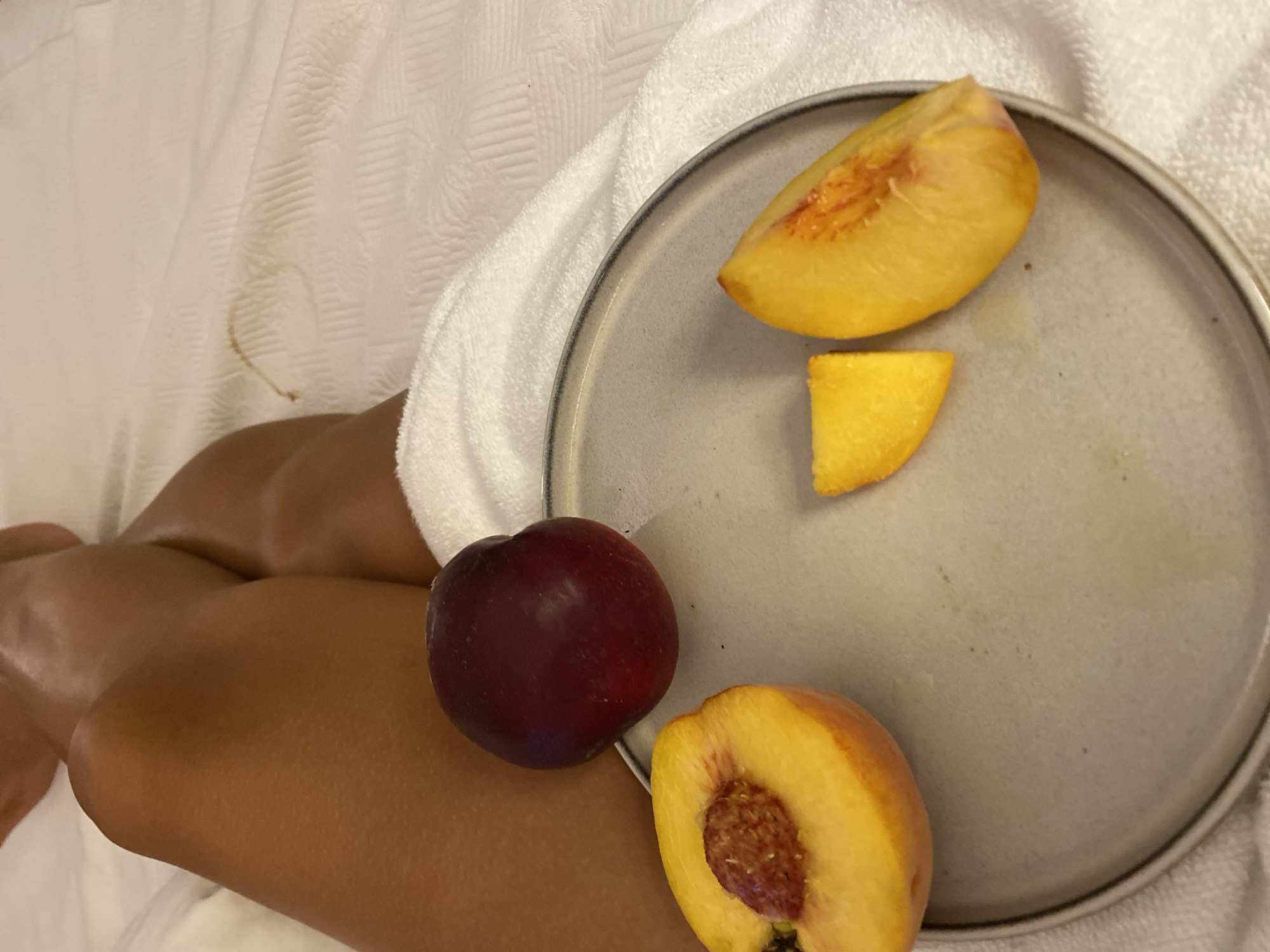 The image displays a close-up view of a person's legs, tanned and resting on a white, textured surface that appears to be a bed or couch. A light-colored towel is draped over the person's lower legs. In the foreground, a gray plate holds pieces of fruit. A whole plum and a halved peach with its pit are visible, along with two sliced pieces of peach, one wedge-shaped and the other a more irregular slice. The background consists of the white textured surface and the person's legs. No specific location within the city of Aksu, Turkey, can be determined from the image itself.