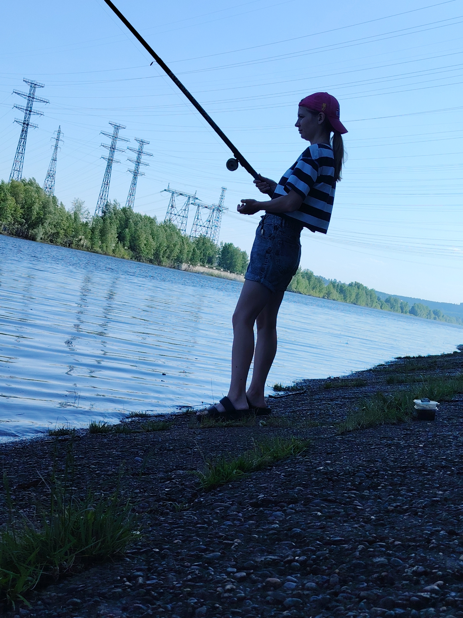 The image shows a young woman fishing on a riverbank. She is wearing a pink baseball cap, a blue and white striped cropped top, and jean shorts. She is holding a fishing rod and appears to be casting or reeling in the line. The river is wide and calm, with trees lining the opposite bank. Electrical pylons are visible in the background. The foreground consists of a rocky shoreline with some grass growing along the edge. The image was most likely taken in Chaikovsky, Russia.