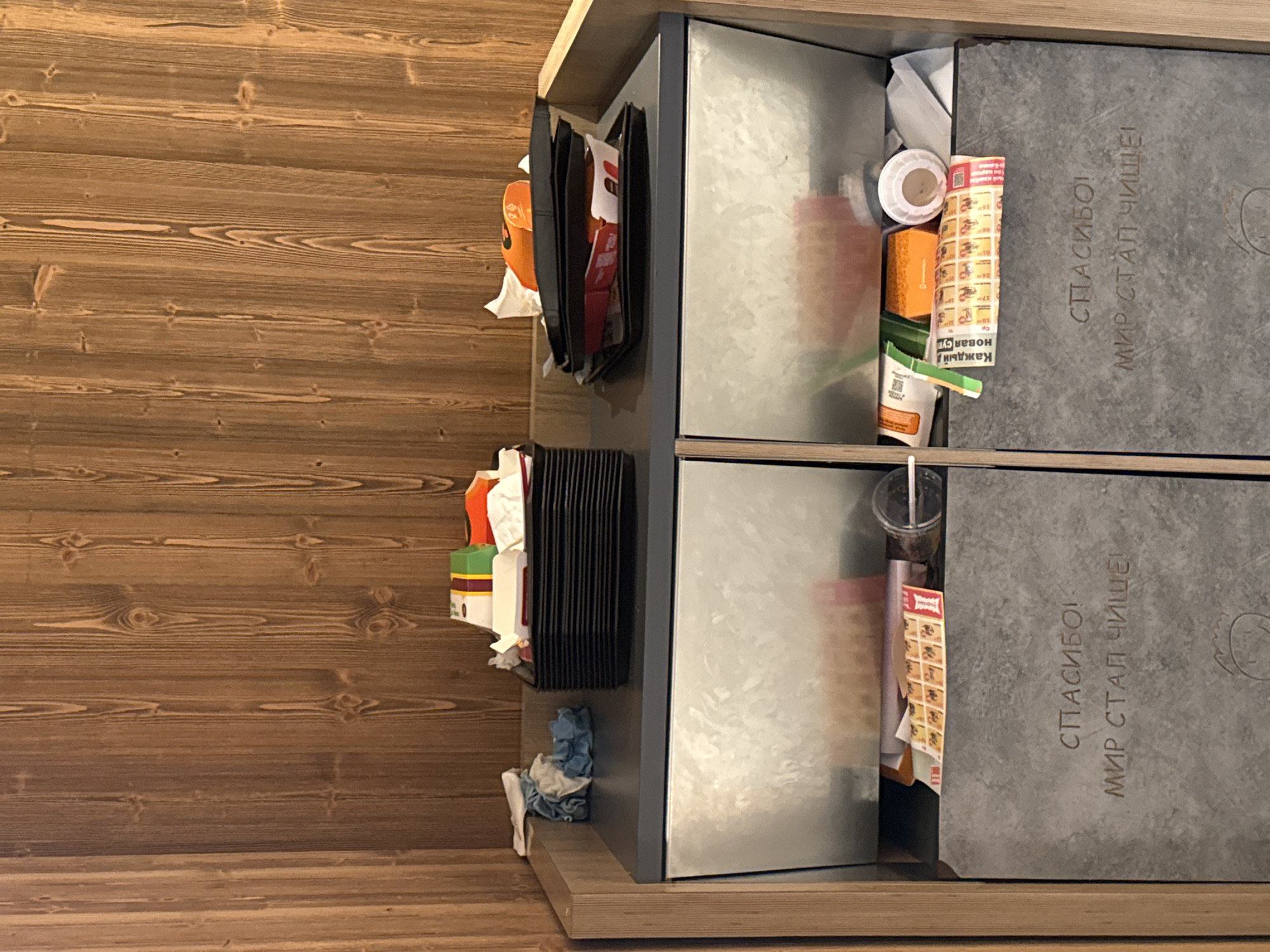 The image depicts a waste sorting bin with two compartments, located in Saint Petersburg, Russia, likely in a public area such as a cafe or fast-food restaurant, given the presence of discarded food containers. The bin has a rustic wooden panel background and a grey stone-like facade at the bottom. Each compartment has a metallic front and contains various items of trash.
The left compartment's top surface is partially visible and shows a stack of black plastic trays, some with white paper liners and crumpled paper. A blue and white crumpled cloth is also present. The interior of the left compartment contains some paper packaging and a clear plastic cup.
The right compartment's top surface is also visible, with more black plastic trays stacked, containing remnants of food packaging, including red and white cardboard boxes and orange plastic. Crumpled white paper and a white plastic lid are also on this surface. The interior of the right compartment appears to hold more paper and plastic waste.
The stone-like facade of the bin has embossed text. On the left compartment's facade, the text reads: "СПАСИБО! МИР СТАЛ ЧИЩЕ!" which translates to "THANK YOU! THE WORLD HAS BECOME CLEANER!". On the right compartment's facade, the same text is embossed: "СПАСИБО! МИР СТАЛ ЧИЩЕ!". Below the text on the right compartment, a simple drawing of a smiling face is also visible.
The overall scene suggests a functional, albeit somewhat messy, waste disposal area designed to encourage recycling or separation of waste. The presence of Russian text on the bin confirms its location in Russia.
