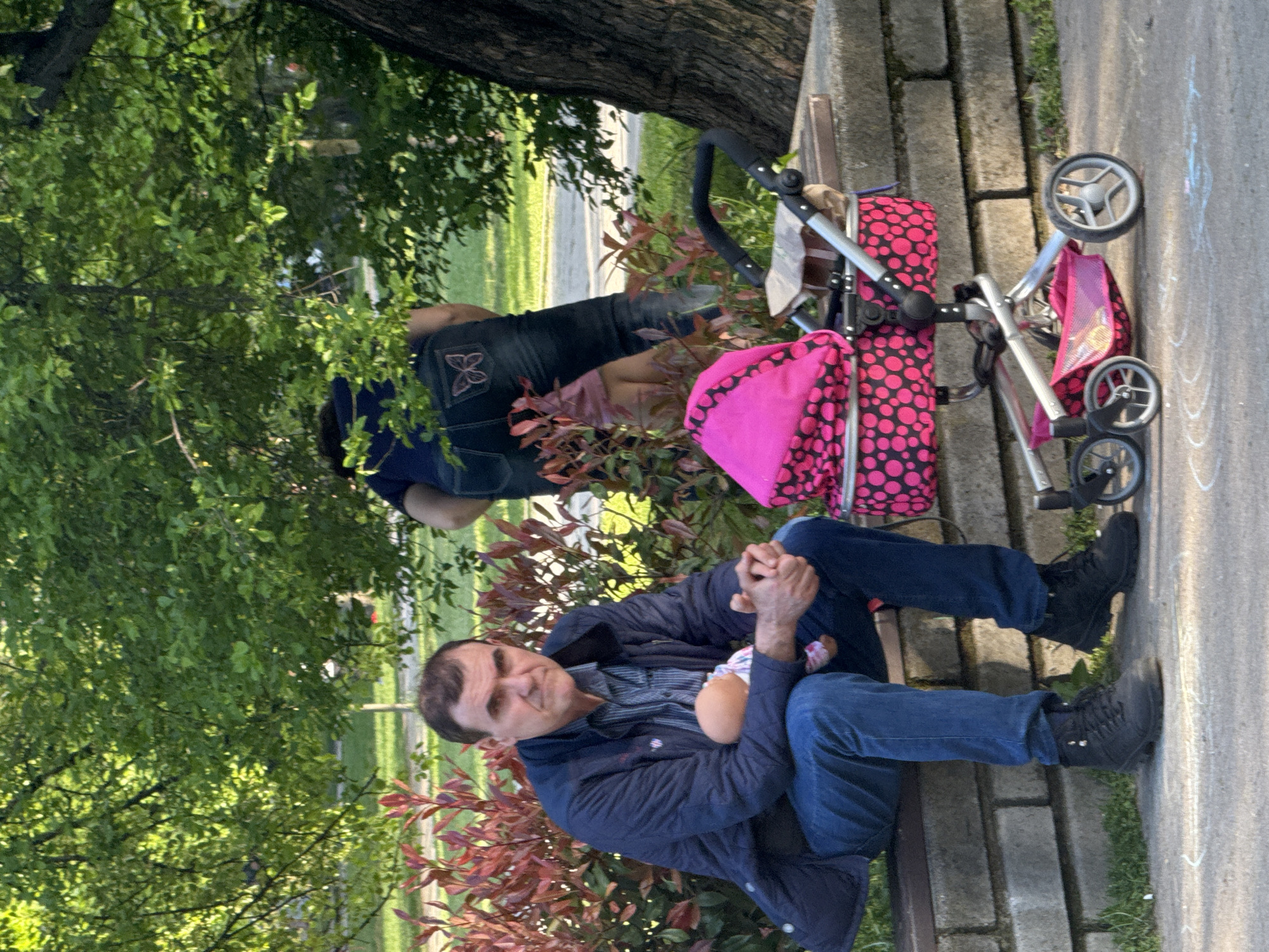 The image shows two people sitting on a bench in a park, potentially in the city of Sofia, Bulgaria. One person is seated upright holding a doll, wearing a blue jacket, striped shirt, jeans, and black boots. The other person is reclining on the bench next to them. Next to them is a stroller with a pink and black polka dot pattern. The bench is situated near a brick wall or path, and there are trees and greenery visible in the background. There is a grassy area with a road nearby.