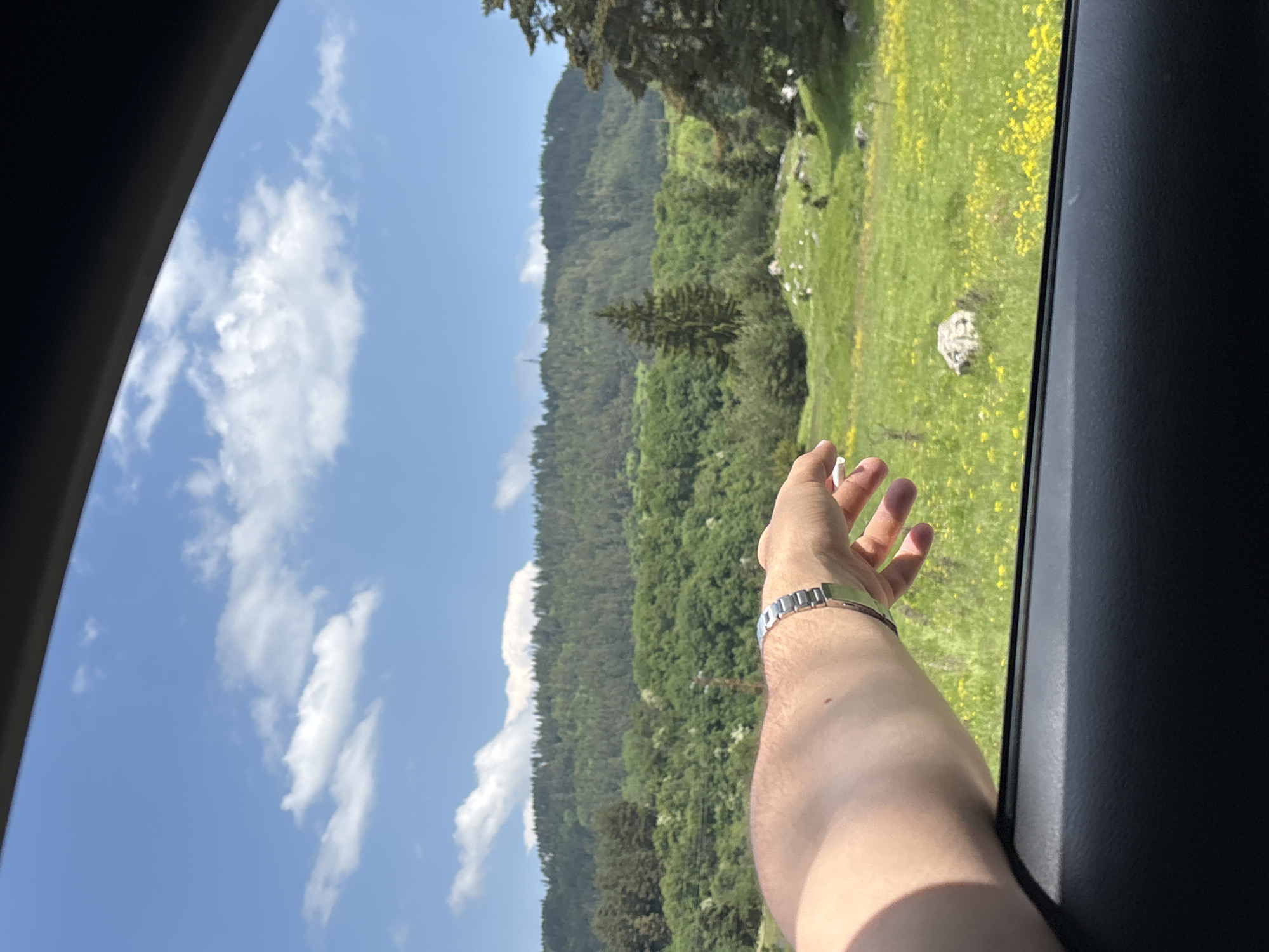 The image presents a sunlit rural landscape observed from the interior of a vehicle, looking out through an open window. The upper third of the frame shows a clear blue sky interspersed with scattered white cumulus clouds. Below the sky, a panoramic view of lush green hills and mountains covered densely with coniferous and deciduous trees dominates the horizon, with a faint utility structure visible on a distant peak. The midground features rolling green meadows, abundantly dotted with small yellow flowers, and scattered light-colored rocks. Several individual evergreen trees and clusters of deciduous trees are interspersed throughout the landscape. In the immediate foreground, a light-skinned human forearm and hand, wearing a metallic link bracelet, extend from the lower-left out of the car window, partially obscuring the lower-left portion of the landscape. The dark grey or black interior of the car's window frame and door panel occupies the bottom section of the image. The scene is brightly illuminated by natural sunlight, casting a shadow from the arm onto the car's interior. The location is specified as Beli Iskar, Bulgaria.