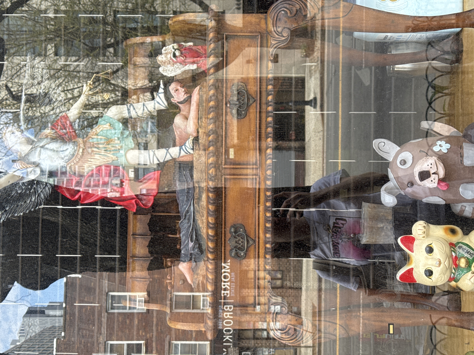 The image shows a store window display likely in the Brooklyn borough of New York City. The display contains several items, including an angel statue holding scales, a partially nude male figurine with bandages around his legs, a wooden door, a stuffed dog toy, and a maneki-neko cat figurine. The reflection in the window shows buildings, a street with lane markings, and a person wearing a t-shirt. The words "Woke Brooklyn" are visible on the display.