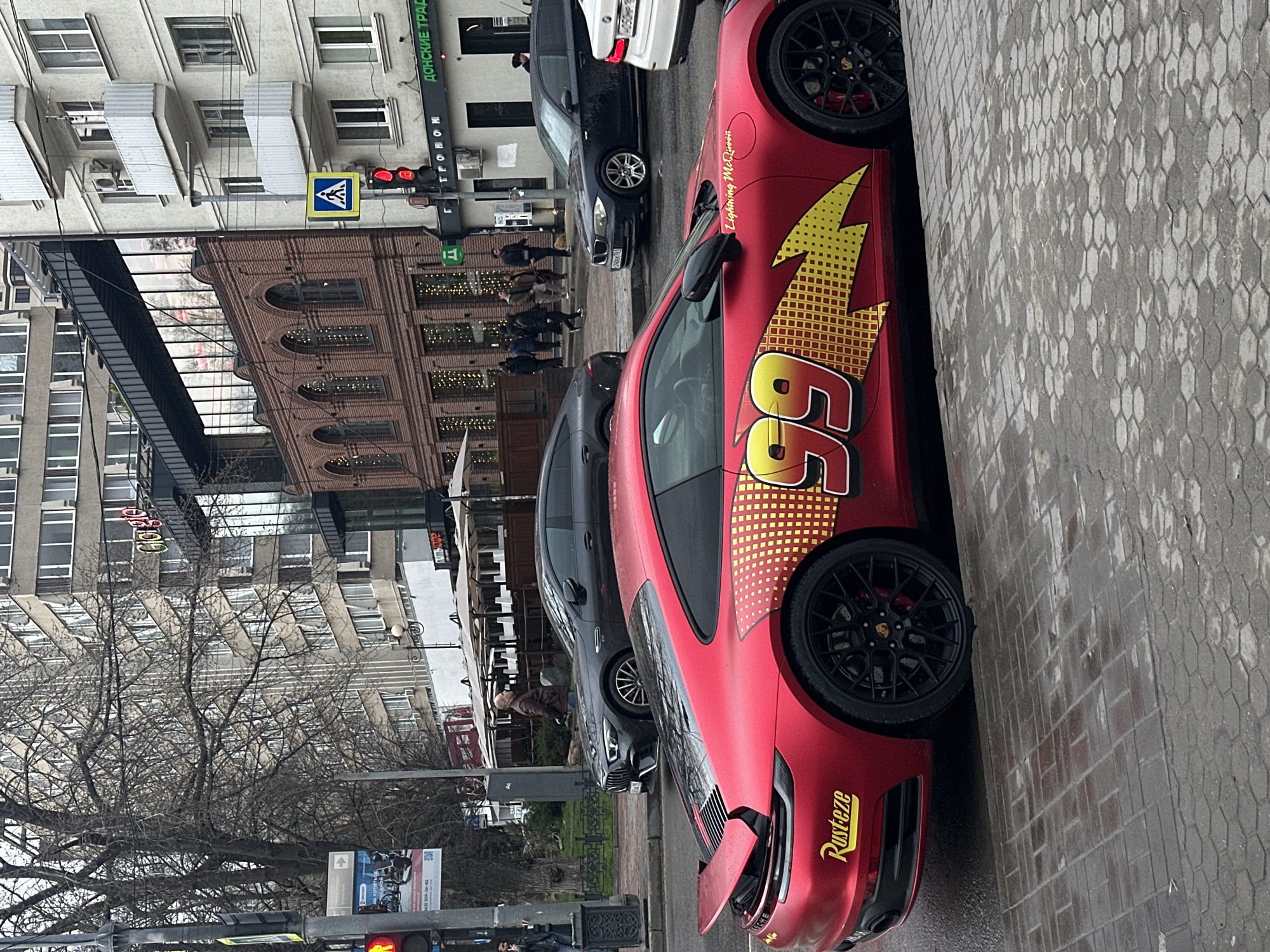 The image depicts a red Porsche 911 adorned with "Lightning McQueen" branding, including the number 95 and "Rusteze" logo, parked on a wet city street. The location appears to be in Rostov-on-Don, Russia, indicated by a sign with Cyrillic text "ДОНСКИЕ ТРАДИЦИИ" visible in the background. Several other cars are parked alongside, and a small group of pedestrians is crossing the street. Buildings with architectural details typical of urban environments line the street. A traffic light is visible, showing a red signal. Bare trees suggest a season with little or no foliage.
