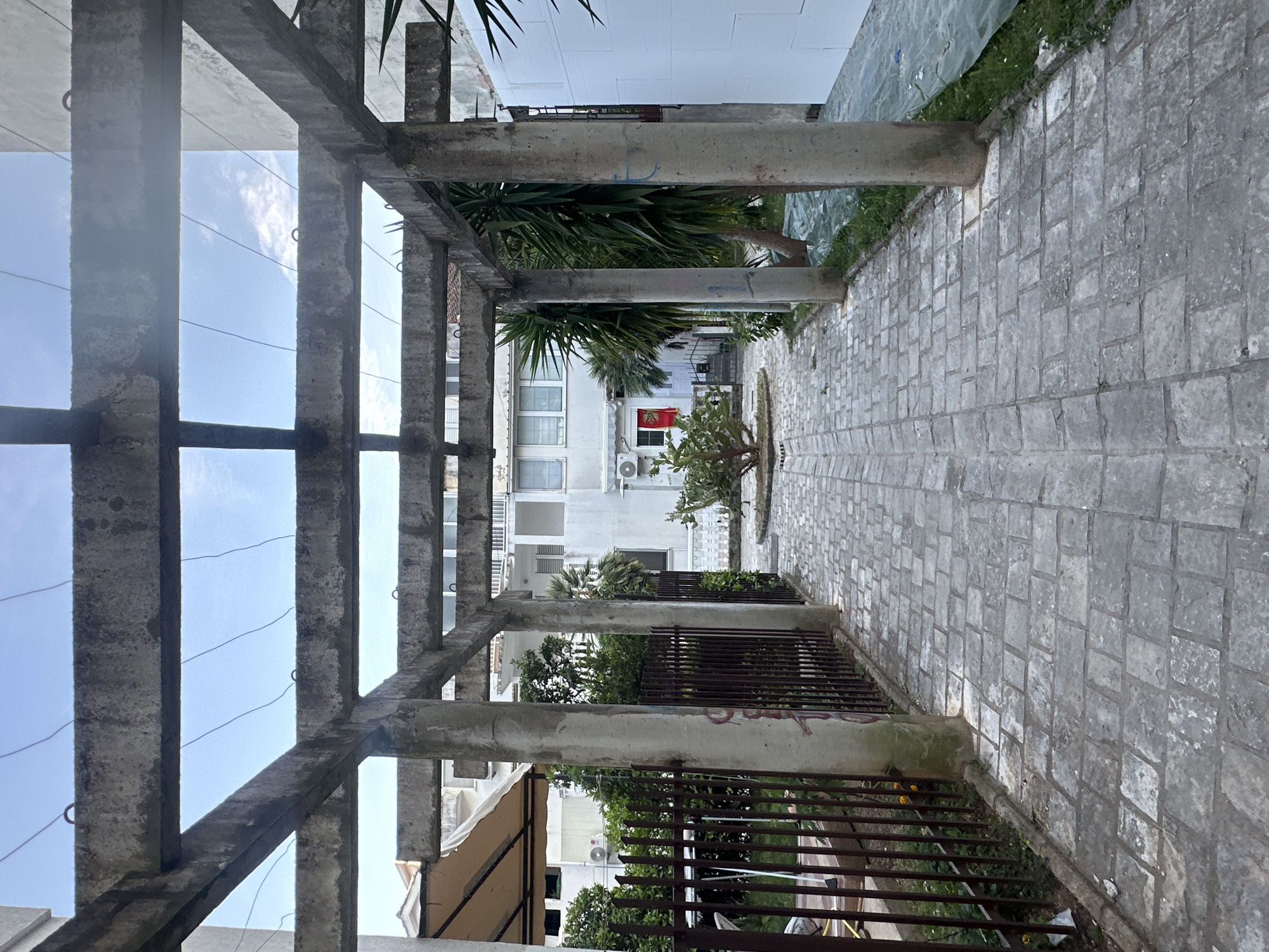 The image shows a paved walkway under a concrete pergola structure. The walkway leads towards a white building with a red flag visible in one of the windows. Metal fencing runs along the right side of the walkway. The location is likely Budva, Montenegro, as suggested by the building style and flag. No people are visible. The sky is visible through the pergola. The concrete structure appears weathered.
