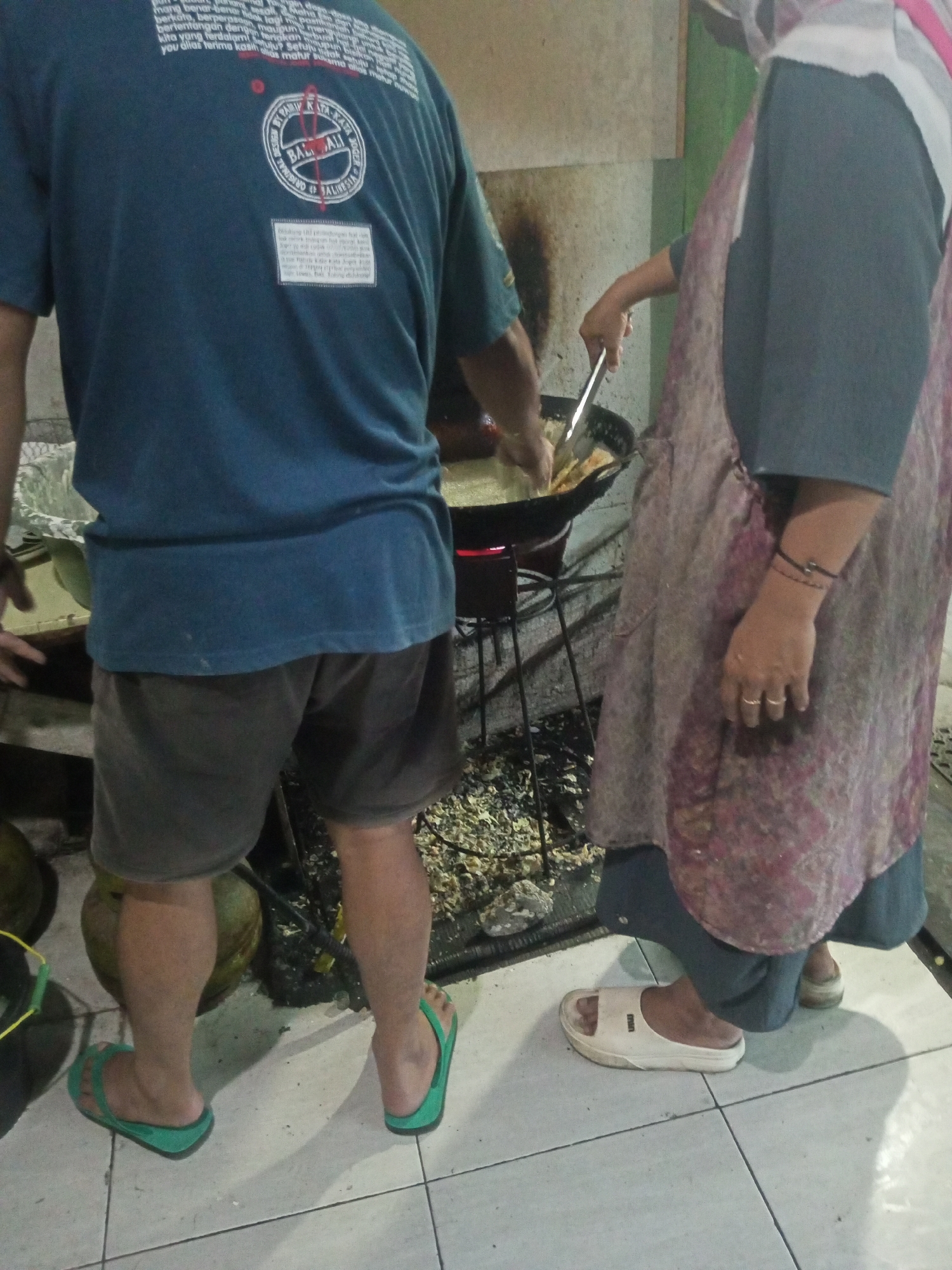 In Andongsili, Indonesia, a person is frying food in a wok over a portable stove. The person on the left, wearing a dark blue t-shirt and brown shorts, is standing near the stove and appears to be observing the cooking process. The person on the right, wearing an apron over a dark blue garment, is actively cooking with a metal utensil in the wok. The environment appears to be an outdoor or semi-outdoor setting with a tiled floor and some scattered debris around the stove. Two large, round objects, possibly coconuts or gourds, are visible to the left of the stove. The lighting is dim, suggesting it might be evening or an indoor area with low illumination.