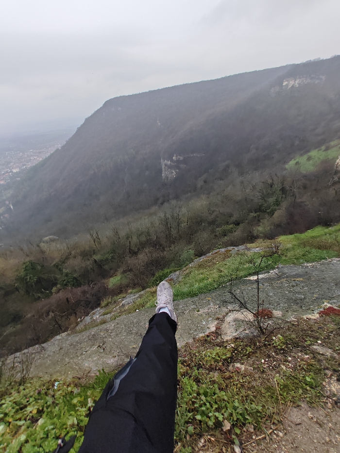 The image depicts a first-person perspective from a high vantage point overlooking the city of Shumen, Bulgaria, and its surrounding landscape on an overcast day. In the immediate foreground, a person's left leg, clad in dark pants and a light grey athletic shoe with a white sole, is extended horizontally over a rocky, vegetated ledge. The leg originates from the lower-left, with the foot positioned roughly in the center-right of the lower frame. The immediate ground beneath the leg consists of grey rock and sparse green and reddish-brown vegetation.
The midground reveals a steep, heavily forested mountainside or large hill, sloping downwards to the left. The trees are predominantly deciduous and appear bare or with minimal foliage, indicating a colder season. Visible amidst the forest are several large, light-colored rock formations or cliff faces. Atmospheric haze or fog partially obscures the details of the distant hills.
In the far background, to the left of the image, a portion of the city of Shumen is visible, characterized by a cluster of buildings and urban infrastructure, nestled at the base of the hills. The sky above is uniformly grey and overcast, providing diffuse, flat lighting to the entire scene.