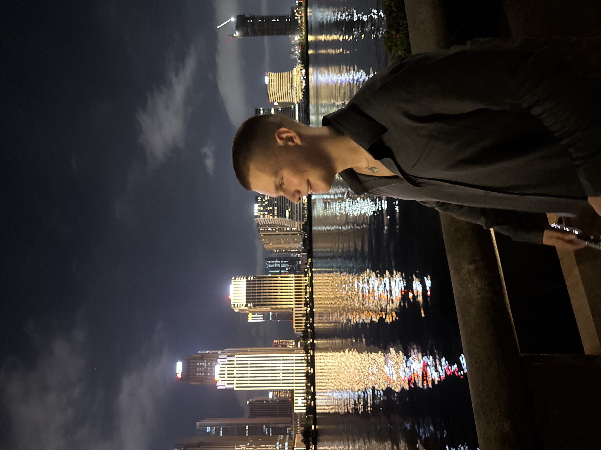 The image displays a nighttime urban landscape in the city of Haizhu District, China. A young adult male, with a short buzz cut and fair skin, is depicted in profile in the lower right quadrant of the frame, smiling and looking towards the right. He wears a dark, long-sleeved collared shirt and a necklace with a small, light-colored pendant. He stands behind a dark, curved stone barrier. Across a wide river that occupies the midground, a modern city skyline is illuminated, with numerous skyscrapers showcasing bright yellow, white, and some red lights that are distinctly reflected on the water's surface. Prominent features of the skyline include buildings with strong vertical light patterns, one with a red corporate logo, and a tall structure under construction on the far right, marked by cranes. The dark night sky is partially covered by scattered clouds, and a faint, singular light source is visible in the upper left portion of the sky.