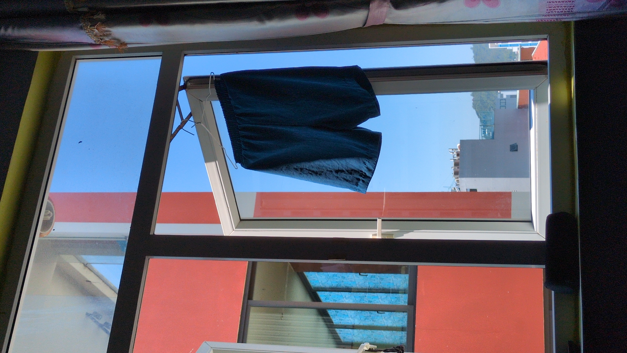 The scene, located in South Quy Nhon Ward, Vietnam, depicts an interior view looking outwards through a multi-pane window under bright daylight. A dark blue pair of shorts, appearing to be made of a lightweight fabric, hangs from a white wire hanger suspended from the top horizontal mullion of a white window frame. The shorts are positioned to the right of the central vertical mullion. To the immediate right of the window, a patterned curtain, white with reddish-pink floral elements and a decorative fringe, partially obscures the view. A dark, cylindrical object rests on the interior windowsill at the bottom.
Beyond the window, a clear, bright blue sky is visible. To the left, a reddish-orange building with dark-framed windows reflecting the blue sky is prominent. A segment of a white vertical structure is positioned between this building and the window frame. In the midground, a light gray building with rooftop structures, possibly HVAC units or water tanks, is visible below the hanging shorts. Further in the background, a green, tree-covered hill or mountain range is discernible under the clear sky, along with other urban buildings, some with reddish-orange rooftops. The interior space from which the photograph is taken appears darker in contrast to the illuminated exterior.