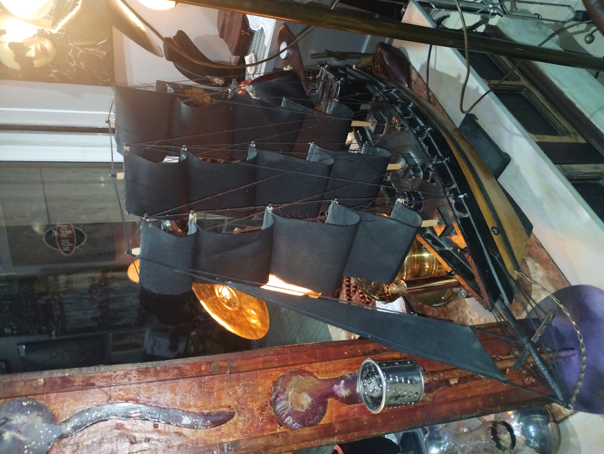 The image shows a close-up, low-angle view of a model ship with black sails and a dark, intricate wooden hull. The ship is positioned on a wooden surface, possibly a table or display stand, which is detailed with what appears to be a faux-worn finish. Several objects are visible around the model ship. To the left, a shiny, golden lamp with a textured surface is illuminated, casting a warm glow. Below the ship, a metallic cylindrical object with perforations is attached to the wooden surface, along with other metallic and decorative elements. In the background, a reflective surface, possibly glass or a mirror, reveals a distorted reflection of the surroundings, including what looks like a shop interior with shelves and various items. A sign with Greek lettering, "ΚΡΙ ΚΟΠ," is visible in the reflection. The lighting is dim and warm, suggesting an indoor setting, possibly at dusk or night. The overall impression is of a display of a decorative or historical artifact in a shop or museum. The location is identified as Perea, Greece.