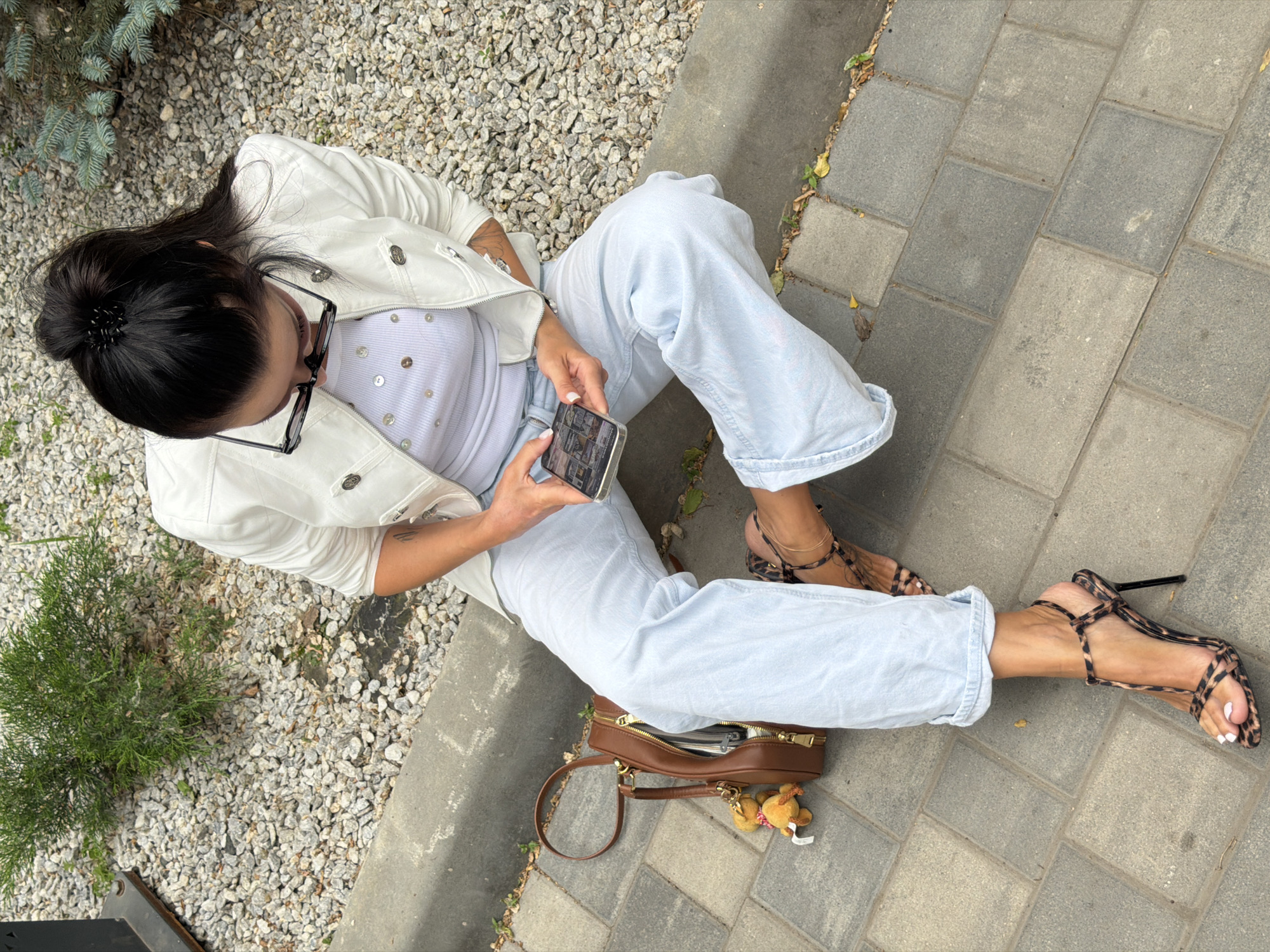A woman is sitting on the ground, looking at her phone. She is wearing light blue jeans, a white jacket, and a white top decorated with iridescent sequins. Her hair is pulled back in a bun with a black clip, and she is wearing black-framed glasses. Her feet are adorned with strappy leopard-print high heels. A brown handbag with a gold zipper and a small teddy bear charm is placed next to her. The setting appears to be outdoors, with a mix of grey gravel and paving stones. A small evergreen bush is visible to her left. The lighting suggests it is daytime. The woman's posture and focus on her phone indicate she is engaged with its content.
