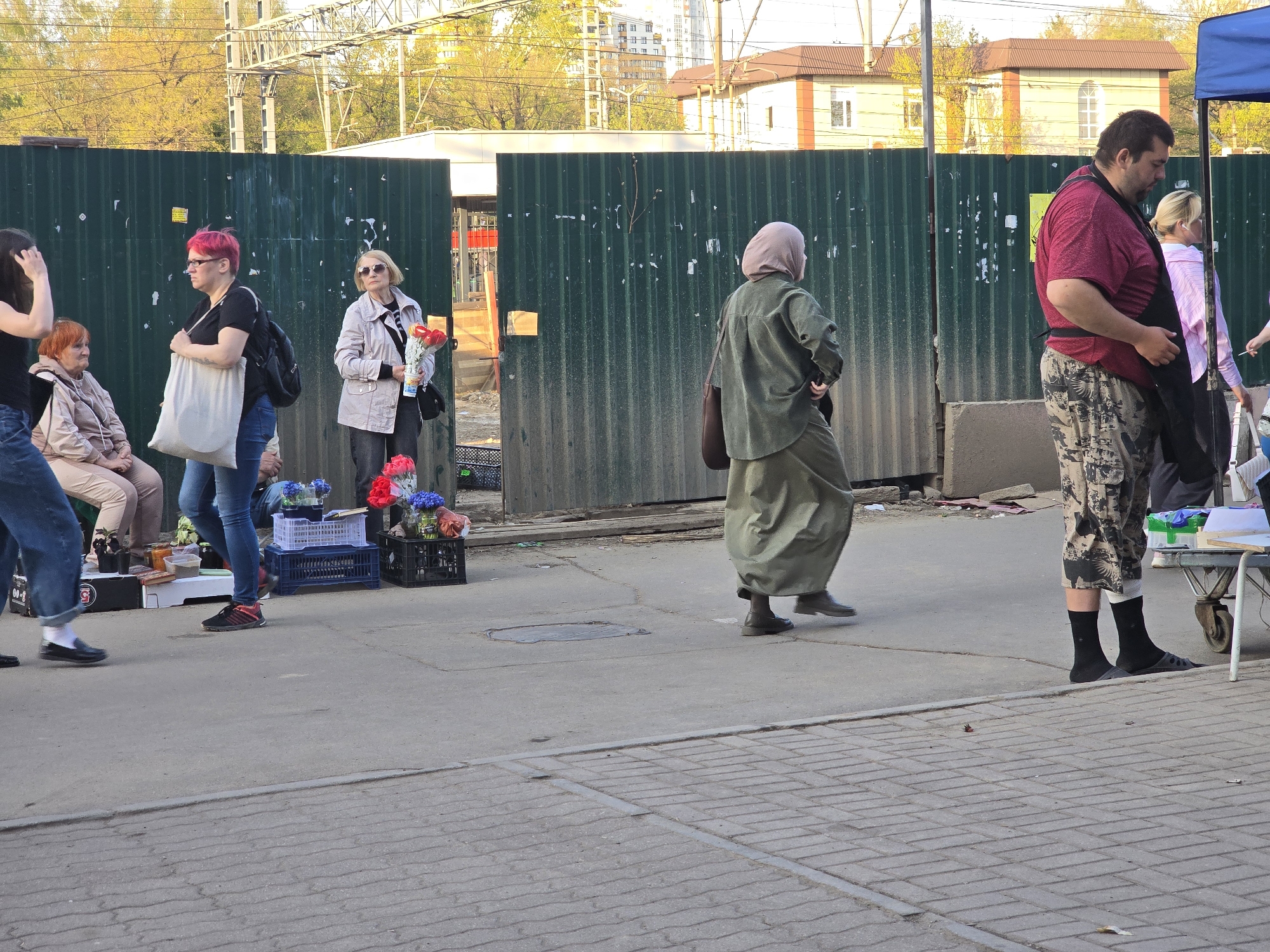 The image depicts an outdoor street scene in Khimki, Russia. A group of people are visible along a sidewalk next to a tall green corrugated metal fence, likely related to construction or a temporary barrier.
People:
- A woman with pink hair, wearing a black shirt and jeans, carrying a white tote bag.
- An older woman wearing sunglasses and a light jacket holds a bouquet of flowers.
- A woman in traditional Muslim dress, including a headscarf, is walking to the right of the frame.
- A man wearing a red shirt, camouflage pants, and black socks with sandals stands near a table.
Objects:
- Crates of flowers for sale are visible near the pink-haired woman.
- A small table is on the right of the image.
- A manhole cover is in the center of the scene.
Environment:
- The sidewalk is made of concrete with patterned bricks near the foreground.
- Buildings are visible in the background.
- Overhead power lines are in the background.