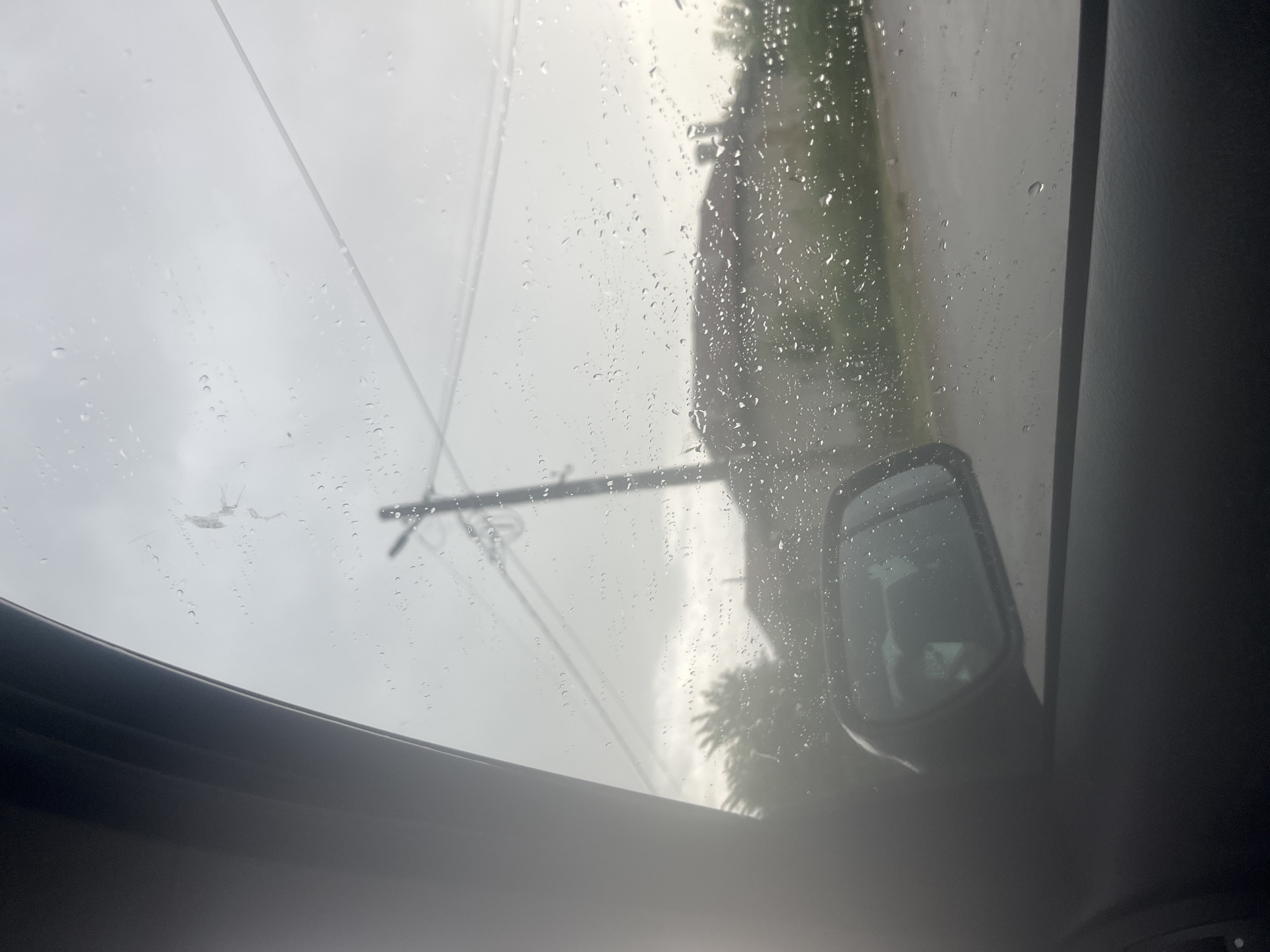 The image is taken from inside a vehicle, looking upwards through a sunroof on a rainy day. Raindrops are visible on the glass. Outside, power lines are visible against an overcast sky. A side mirror of the vehicle is also visible. The general landscape seems to include some greenery, possibly trees and foliage. The location, Byala Cherkva, Bulgaria, cannot be confirmed solely from the visual information.