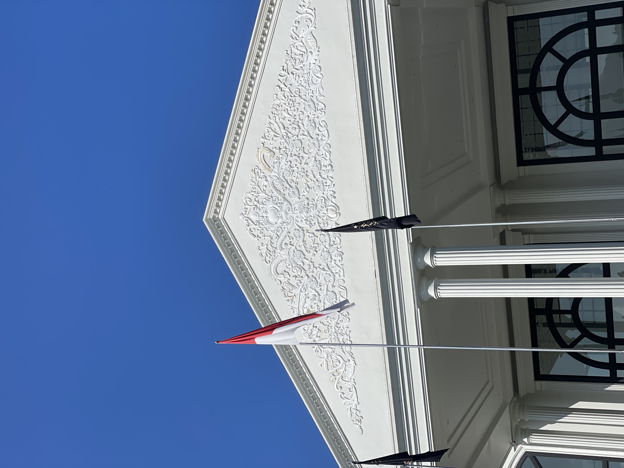 The image displays the upper portion of a white neoclassical-style building against a clear, vibrant blue sky. The sky occupies the upper half of the frame, devoid of clouds. The building's prominent feature is a white triangular pediment adorned with intricate white bas-relief floral and acanthus scrollwork patterns. A subtle, light gold-colored accent is visible within the relief on the left side of the pediment.
Three flags are visible:
1. Centered on the facade is a red and white bicolour flag, consistent with the national flag of Indonesia, attached to a silver flagpole and hanging vertically.
2. To the left of the Indonesian flag, a black flag with partially visible white or light-colored text (appearing as "A A A") is mounted.
3. To the right of the Indonesian flag, another similar black flag with white or light-colored text is also mounted.
Below the pediment, two white fluted columns are visible, supporting an overhang. The underside of this overhang features a white paneled ceiling. At the bottom of the frame, portions of large dark-framed windows or glass doors with arched tops are visible, reflecting ambient light. No individuals are present in the scene. The setting is in Kayuambon, Indonesia.