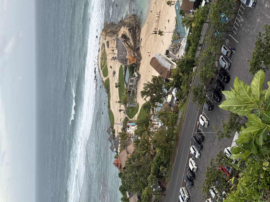 An elevated, wide-angle view depicts the coastal area of Melasti Beach in Ungasan, Indonesia, under an overcast sky. The image is structured from foreground to background:
The immediate foreground features dense green foliage, partially obscuring a paved parking lot laid with grey interlocking bricks, which contains several parked cars of various colors (white, black, red, silver).
A two-lane asphalt road runs horizontally across the lower midground. Flanking the road are additional parking areas, densely filled with dozens of parked cars and vans, separated by small trees.
Immediately below the road, a resort complex is visible, characterized by numerous buildings with traditional red-tiled and thatched roofs, several blue-water swimming pools (one featuring slides), and abundant tropical vegetation, primarily tall palm trees. Small groups of people are faintly discernible near the pools and buildings.
The midground transitions to a sandy beach where numerous individuals and small groups of people are scattered, some near the water's edge. A distinctive, large, dark-wood structure resembling a multi-masted sailing ship is positioned on the sand. Adjacent to this ship structure, a rocky breakwater extends into the ocean, topped with green grass and paths, with a few people visible on it.
The background consists of the Indian Ocean, exhibiting white-capped waves breaking both near the shore and further out, beneath a uniform grey, cloudy sky.