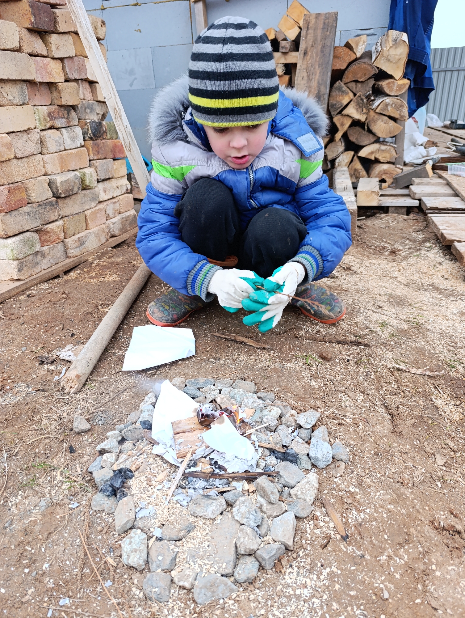 A young boy, dressed in winter attire, is crouched outdoors, intently observing a small, smoldering fire. He wears a blue and gray hooded winter jacket with a fur trim, black pants, a striped black, gray, and yellow beanie hat, and white and green gloves. His patterned shoes are visible as he kneels on the dirt and wood shavings.
The small fire is contained within a circular arrangement of irregular gray rocks, with paper and small pieces of wood burning or charred. The boy's gloved hands are clasped together, holding a thin stick, and his expression is one of deep concentration or curiosity as he looks at the flames.
The setting appears to be a rustic or outdoor work area, possibly in Chaikovsky, Russia, during cool, overcast daytime weather. To the left, a tall stack of reddish-brown bricks is visible. Behind the boy, to the right, are neatly stacked logs of firewood and some wooden planks. A light-colored fence and what looks like a concrete block structure are in the background. A long wooden pole lies on the ground near the boy's left knee. The ground is a mix of dirt, wood chips, and scattered debris. No visible text is present in the image.