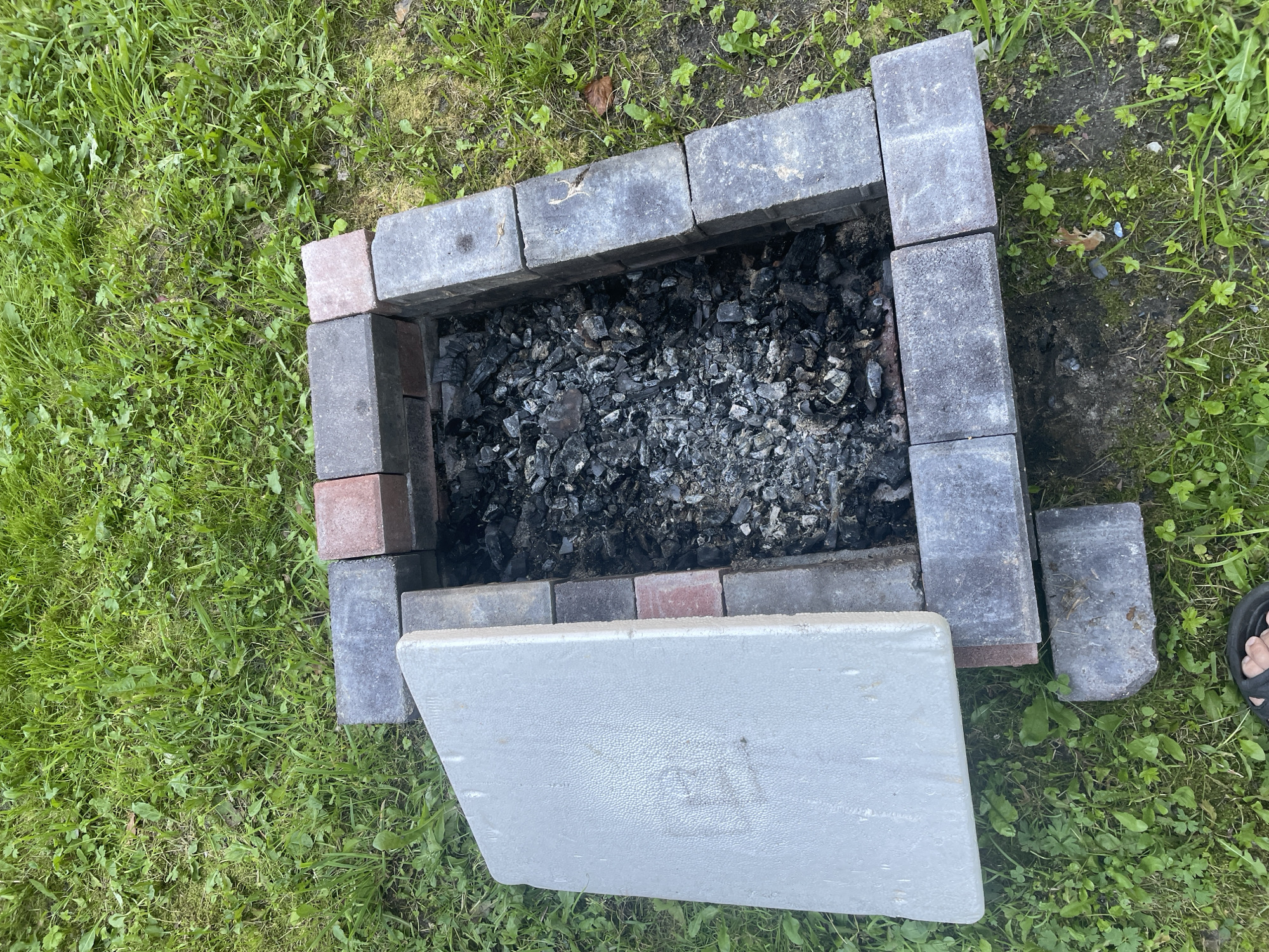 A fire pit constructed of bricks sits in a grassy area, likely in a yard. The fire pit contains a substantial amount of grey ash and charcoal. One side of the fire pit is partially covered by a light grey, rectangular lid with a textured surface. The lid is ajar, revealing the contents of the pit. The surrounding environment consists of green grass with some patches of dirt and a few fallen leaves. A person's foot wearing a black sandal is visible in the bottom left corner of the image, suggesting the photo was taken from a low angle. The scene appears to be outdoors, possibly a backyard or a park. There is no information to ascertain the location in the city of Vidnoye, Russia.