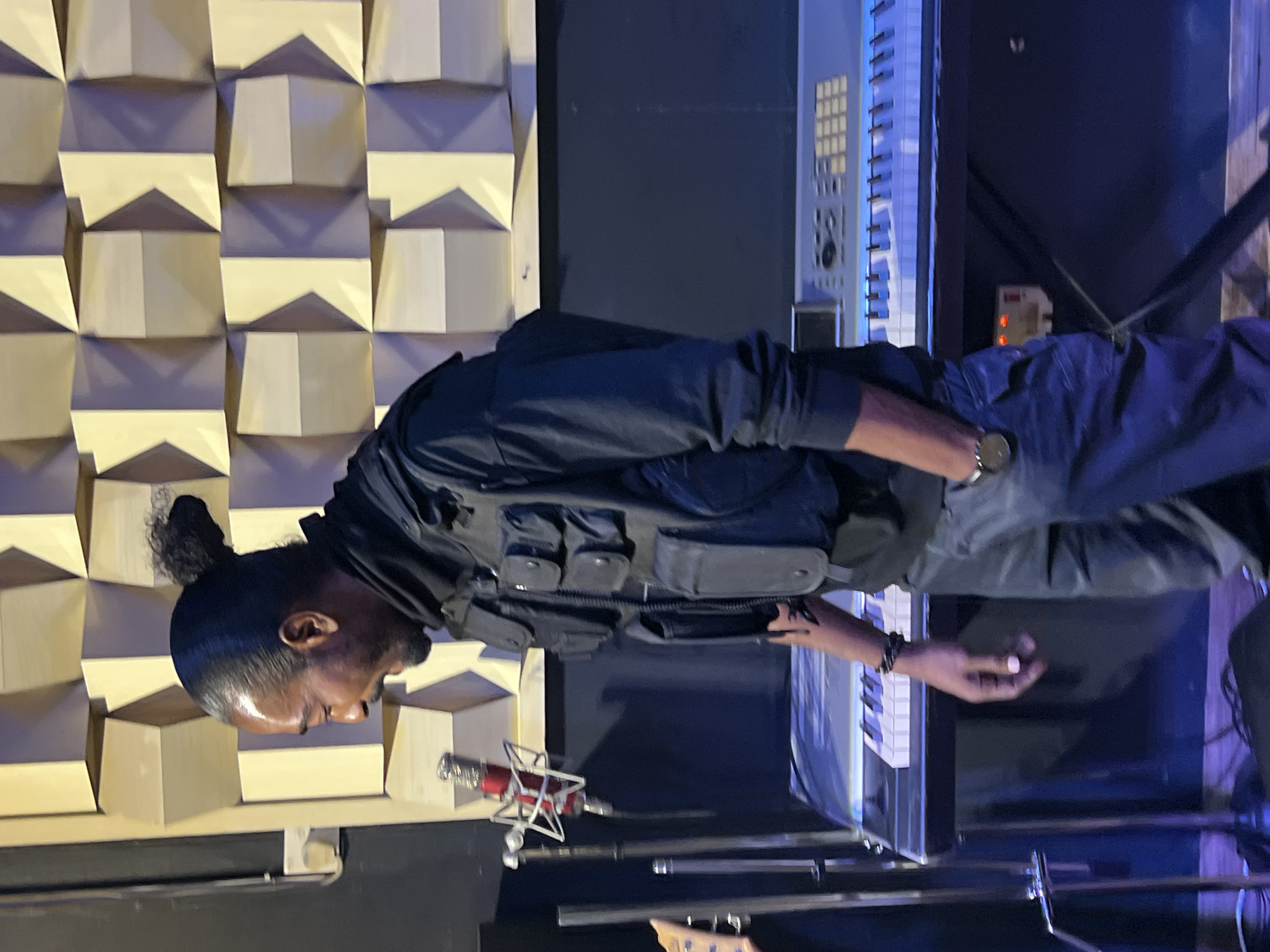 The image shows a Black man with his hair tied in a bun standing in what appears to be a recording studio. He is wearing a black tactical vest over a black turtleneck and gray pants. He is looking to his left, with his mouth open as if speaking or singing. In the background, there is a microphone on a stand, a keyboard, and a wall with textured acoustic panels. The lighting is dim, with blue and purple hues. The man's right hand is resting in his pocket, and his left hand is visible near the microphone. The environment suggests a music production or performance space. The overall scene is focused on the individual and his immediate surroundings. The provided context about the city of Jos, Nigeria, is not visually discernible from the image itself.