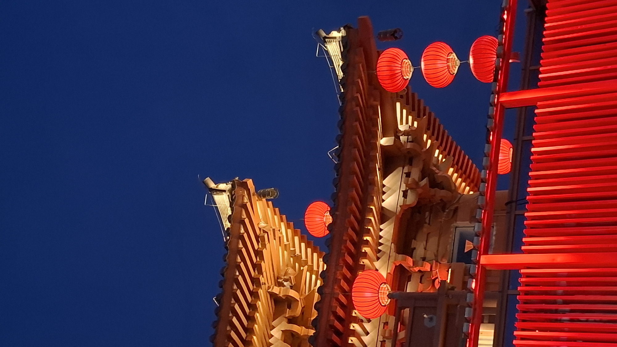 At night in the Yanta District of China, two ornate, multi-tiered buildings stand illuminated against a deep blue sky. The building on the left is warmly lit in orange hues, while the building on the right features bright red accents, including rows of horizontal slats and traditional red lanterns. The lanterns are spherical and appear to be lit from within. The architecture is intricate and detailed, with numerous carved elements and tiered roofs. The scene is captured from a low angle, emphasizing the height and grandeur of the structures.