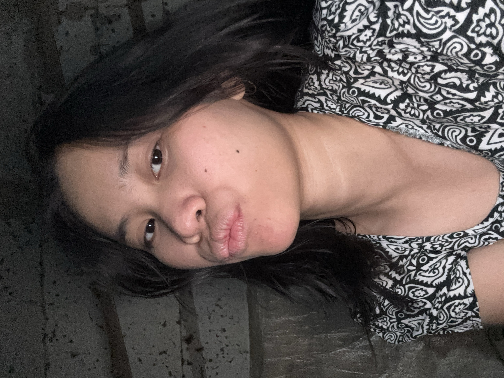 A young woman with dark hair and a black and white patterned shirt puckers her lips for a selfie. She is looking at the camera and her face is partially obscured by her hair. The background appears to be a dimly lit, textured wall. This image was taken in East Jakarta, Indonesia.