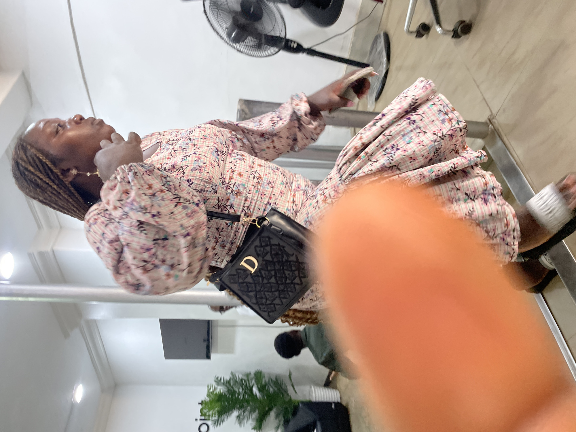 A woman is indoors, appearing to be in a waiting area or salon. She has brown hair braided with lighter strands, wearing dangle earrings, and is wearing a long-sleeved floral patterned dress with a black Dior purse over her shoulder. She is holding a phone in her hand. There is an electric fan mounted on the wall behind her. In the background, a black television, a plant and a person are visible. Part of a finger is obscuring the lower right of the image. The location is likely in Jos, Nigeria, based on the prompt.