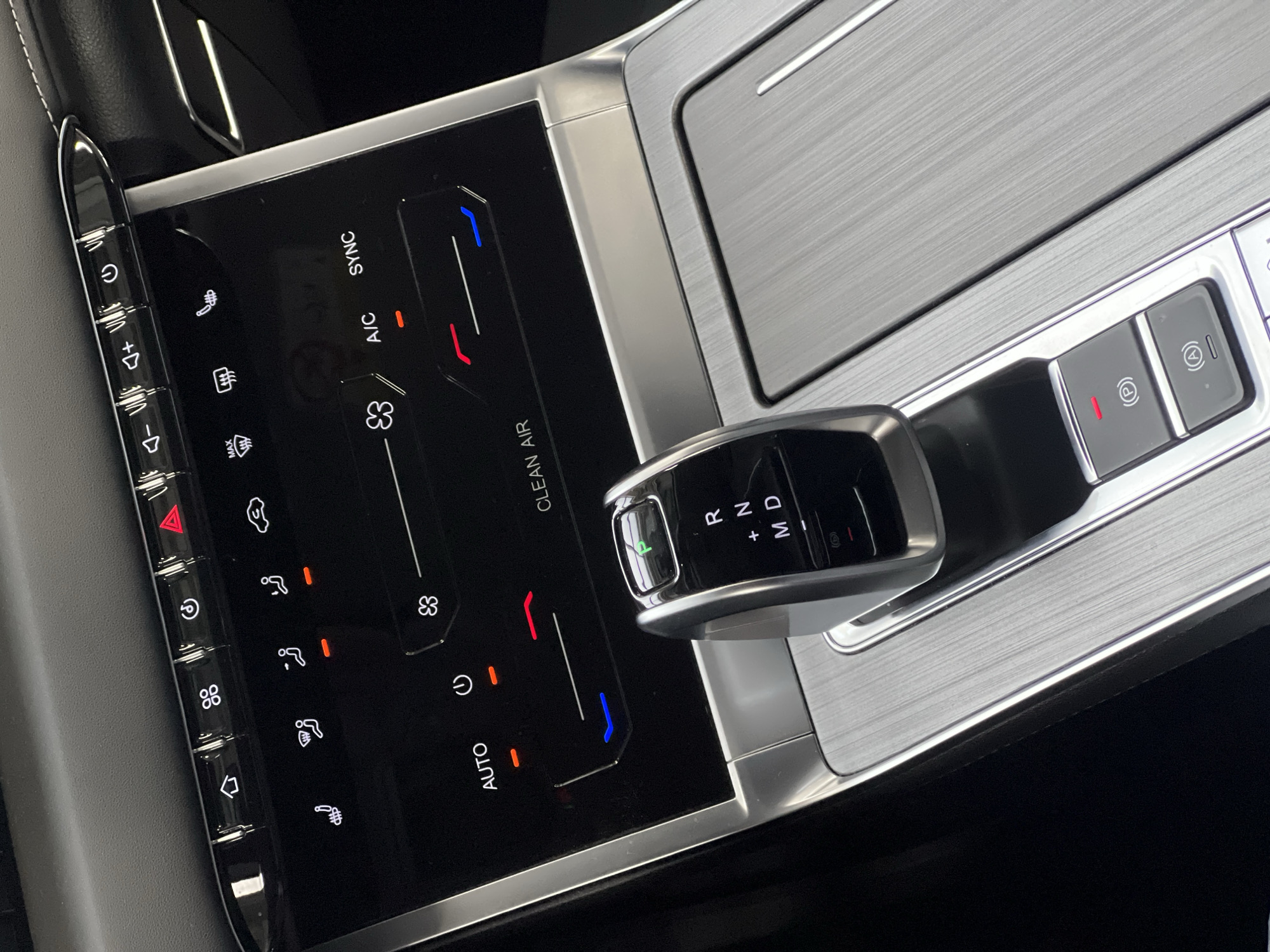 The image displays the interior of a modern vehicle, focusing on the center console and dashboard controls. The perspective is from slightly above and forward, looking down into the cabin.
At the top of the frame, a dark, textured dashboard surface with white stitching is visible. Below this is a glossy black touch-sensitive screen integrated into the dashboard, serving as the climate control and infotainment interface. Above the screen, a row of five distinct physical buttons with chrome surrounds is present. From left to right, these include a home icon, a dual-fan/vent icon, a clock or recirculation icon, a red illuminated hazard light triangle, and a heated seat/defrost icon. To the right of these, a separate cluster of smaller physical buttons controls audio volume (down and up) and power for the infotainment system, accompanied by another heated seat/defrost icon.
The main screen displays various climate control settings. On the upper portion, illuminated icons indicate functions such as heated seats (one showing as active), front and rear defrosters, and air circulation. The words "CLEAN AIR" are visible at the bottom right of the screen section. Below this, two horizontal sliders represent temperature or fan speed, with a fan icon above them. Both sliders show red illuminated indicators, while a blue line is present under the right-hand slider. Text labels "AUTO", "A/C", and "SYNC" are also displayed on the screen.
In the center of the console is a modern electronic gear selector, finished in silver and black. It clearly displays "P," "R," "N," "+," "M," and "D" for various drive modes. The "P" (Park) indicator is illuminated in green.
The area surrounding the gear selector and extending to the right features a brushed metallic trim, appearing to be aluminum, creating a sophisticated aesthetic. Below the gear selector, two rectangular black buttons with chrome surrounds are positioned. The top button, marked with a parking brake symbol "(P)", has a red indicator light illuminated. The bottom button, marked with an auto hold symbol "(A)" encircled, is not illuminated. To the right, the brushed metallic surface continues, suggesting a covered storage compartment or wireless charging pad area.
No individuals are visible in the scene. The image provides no identifiable geographical features or landmarks that would indicate the specific location within the city of Izhevsk, Russia, or any other location.
