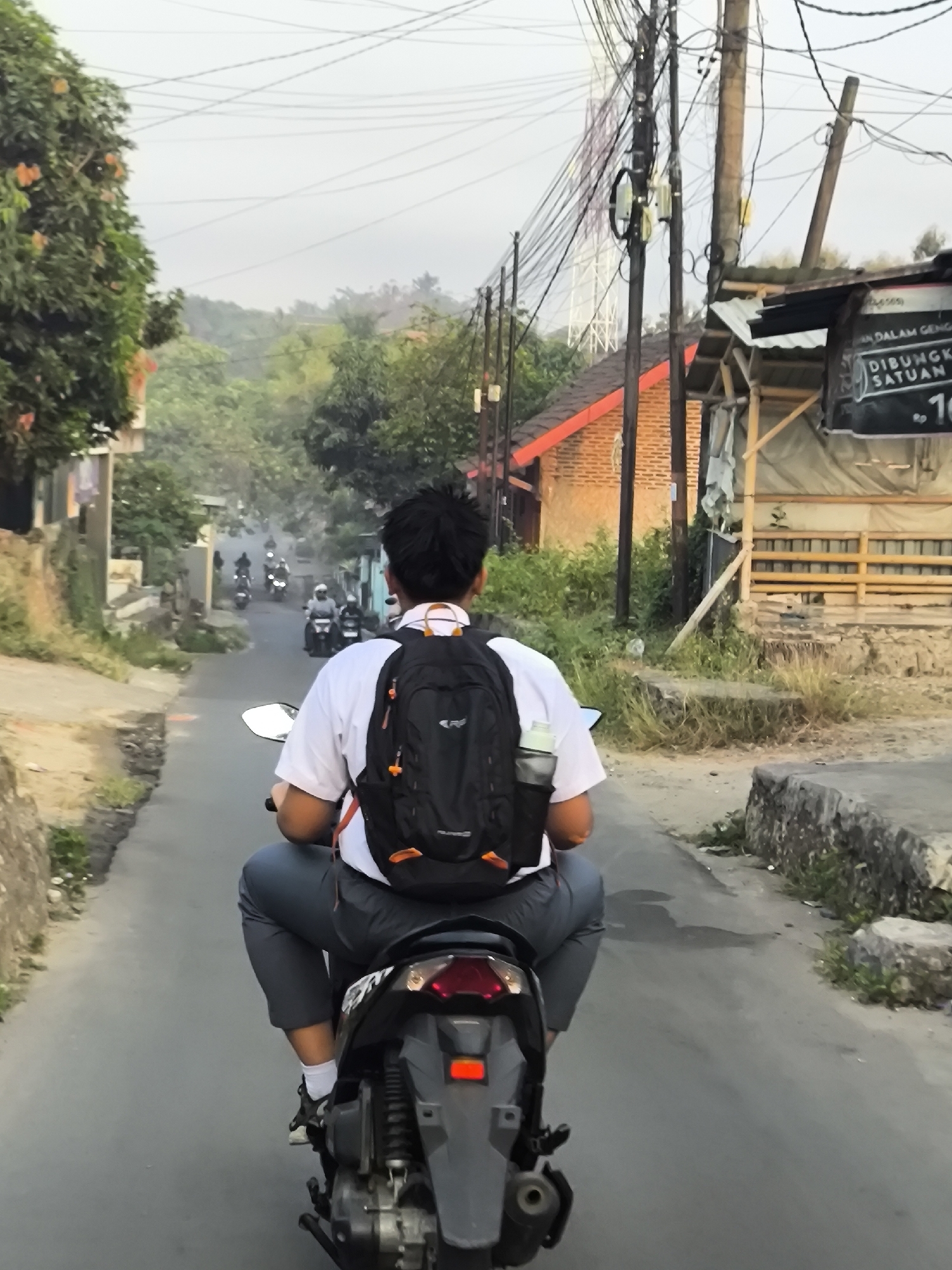 The image depicts a young person wearing a white shirt, gray pants, white socks, and black shoes, riding a black motorcycle with a black backpack. They are viewed from behind, traveling down a paved road. The environment appears to be a residential or semi-rural area with buildings and vegetation on either side of the road. In the background, several other motorcycles are visible, suggesting traffic. Electrical wires are prominently displayed overhead. A sign with Indonesian text is visible on a structure to the right. The sky is overcast, and the overall lighting suggests it is either early morning or late afternoon. Based on the context provided, this scene is located in Cilegon, Indonesia.
