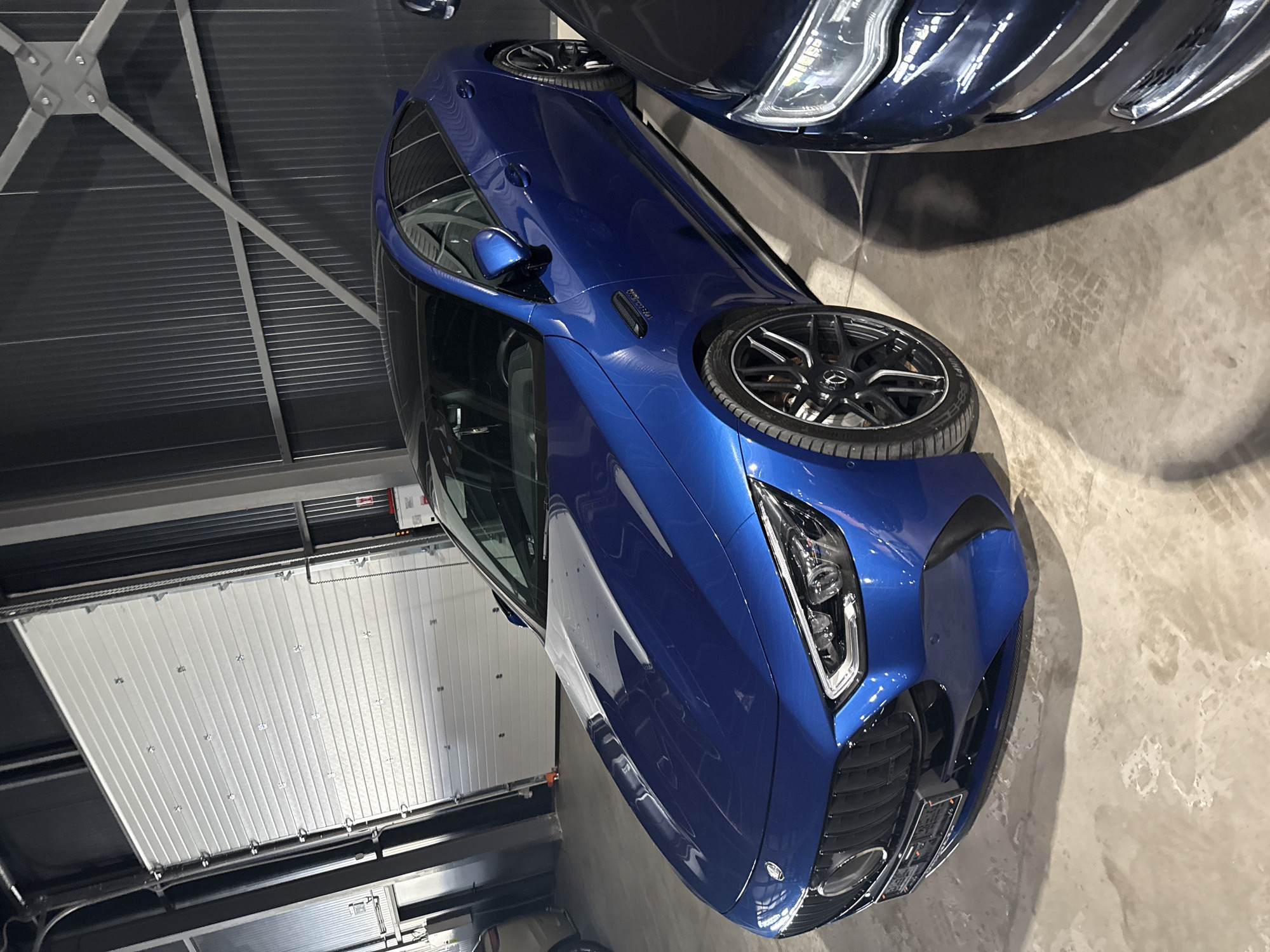 A bright blue Mercedes-AMG GT 63 S E Performance car is parked in a garage with a large roll-up door in the background. The car is positioned at an angle, with its front left side closer to the viewer. The car's sleek, aerodynamic design is evident, with its distinctive grille, LED headlights, and black alloy wheels. The interior of the garage is industrial, with corrugated metal walls and exposed beams. Another dark-colored car is partially visible on the right side of the frame. The floor is concrete and appears slightly wet or stained. There are no people visible in the image. The location is Plovdiv, Bulgaria.