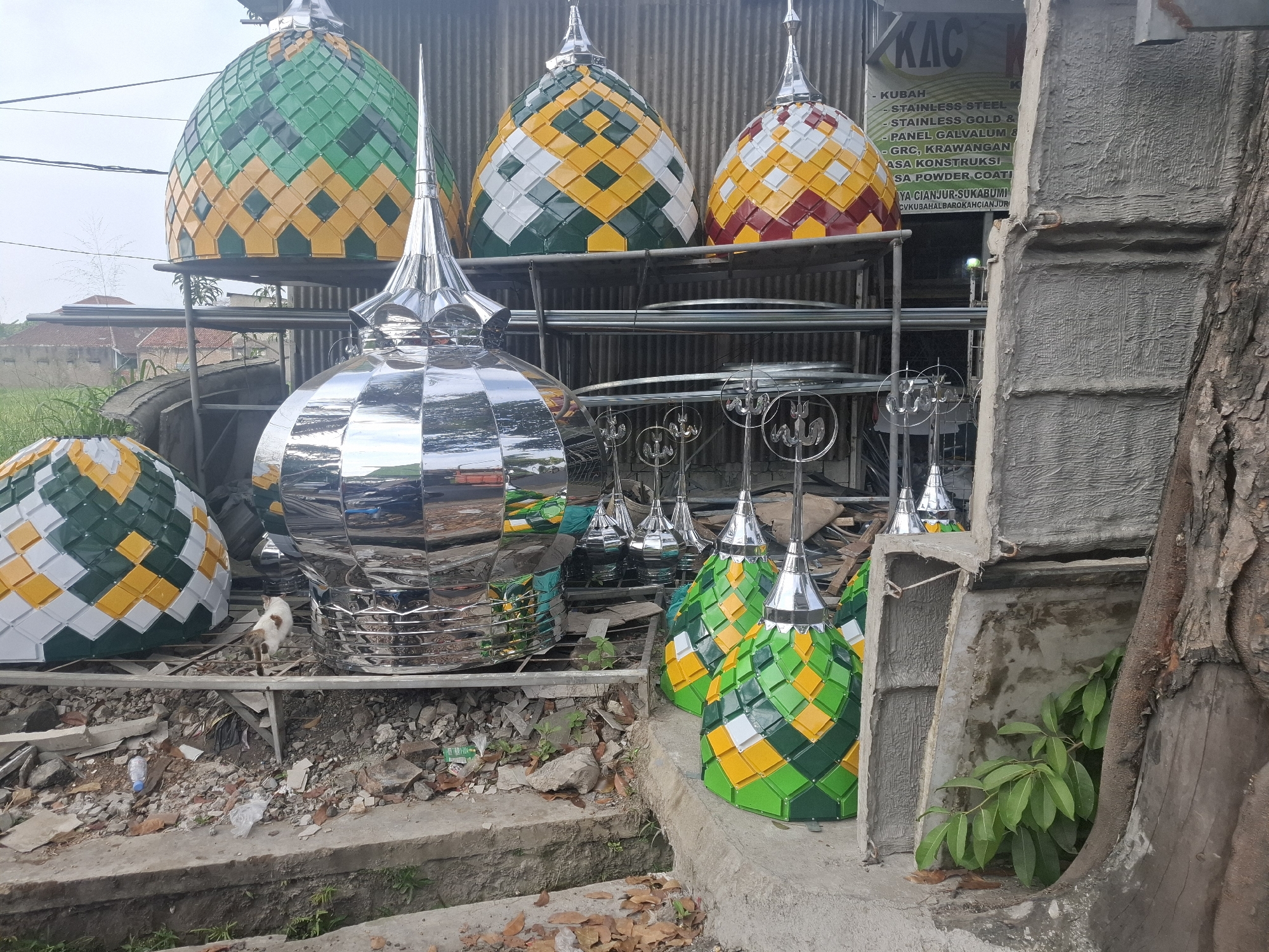 This image depicts a lot filled with various types of mosque domes, likely at a manufacturing or sales site in Rancagoong, Indonesia. In the foreground, several large, shiny, silver-colored domes are prominently displayed. To the left, a smaller dome with a pattern of green, yellow, and white squares is visible. Behind these, on metal racks, are more domes, some with similar geometric patterns in green, yellow, and white, and others in red, yellow, and white. One of these domes on the rack appears to have a slightly different pattern of green, yellow, and white squares. In the background, more racks hold what appear to be smaller, silver finials or decorative elements. To the right, a large, rough-textured concrete structure and a thick tree trunk frame the scene. The ground is covered with debris, including broken concrete, leaves, and discarded plastic bottles, suggesting an outdoor, possibly unfinished or industrial, setting. A white and tan cat is walking on the metal racks among the domes in the mid-ground. The sky is overcast. A sign with text, partially obscured, is visible on a wall behind some of the domes.