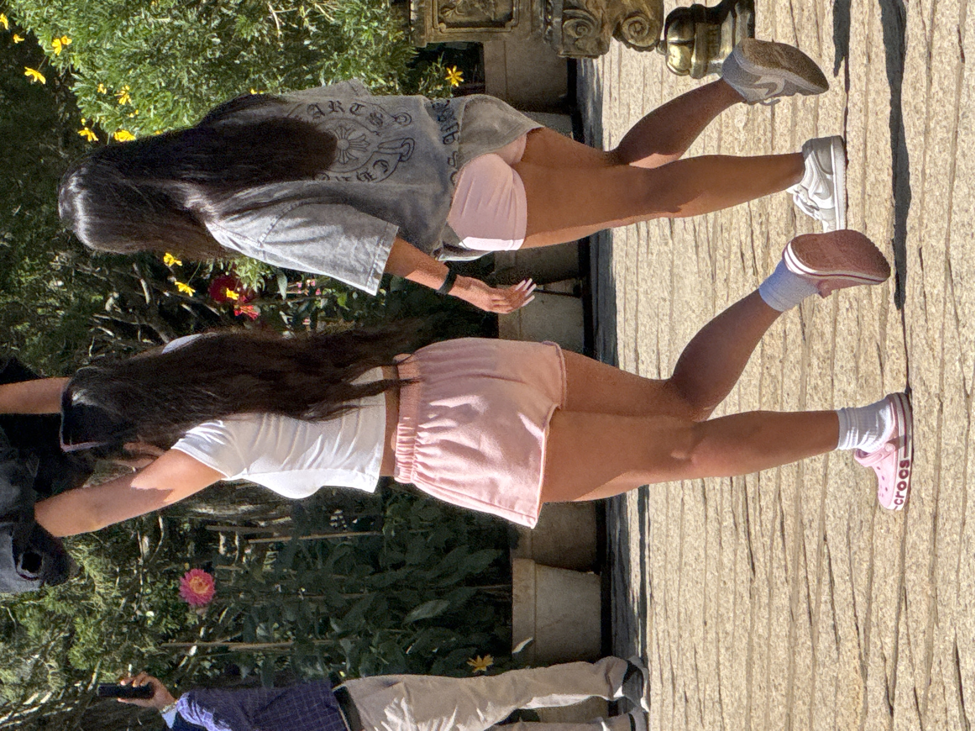 The image shows two young women walking on a cobblestone path in an outdoor setting, likely a garden or park in Da Lat, Vietnam. The woman on the left is wearing a white t-shirt and pink shorts, and pink Crocs with white socks. She is holding a black object above her head. The woman on the right is wearing a grey t-shirt and pink shorts, and white sneakers. She is walking slightly ahead of the first woman. In the background, there are potted plants and yellow and red flowers. The overall impression is of a sunny day in a vibrant, natural environment.