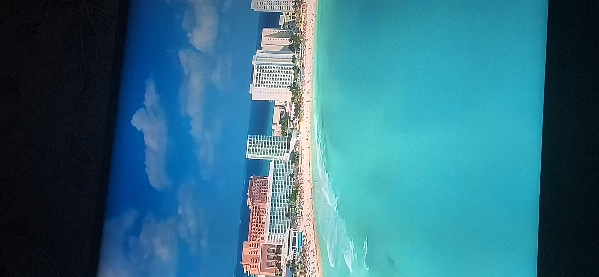 The image depicts a coastal scene with several large, modern hotel buildings situated on a sandy beach. The ocean in the foreground is a vibrant turquoise color with gentle waves approaching the shore. The beach is populated with numerous people, some sunbathing and others walking along the water's edge. The hotels are tall structures with many windows, typical of resort destinations. The sky above is blue with scattered white clouds. This scene does not appear to be located in Bila Tserkva, Ukraine, as Bila Tserkva is an inland city without a coastline.